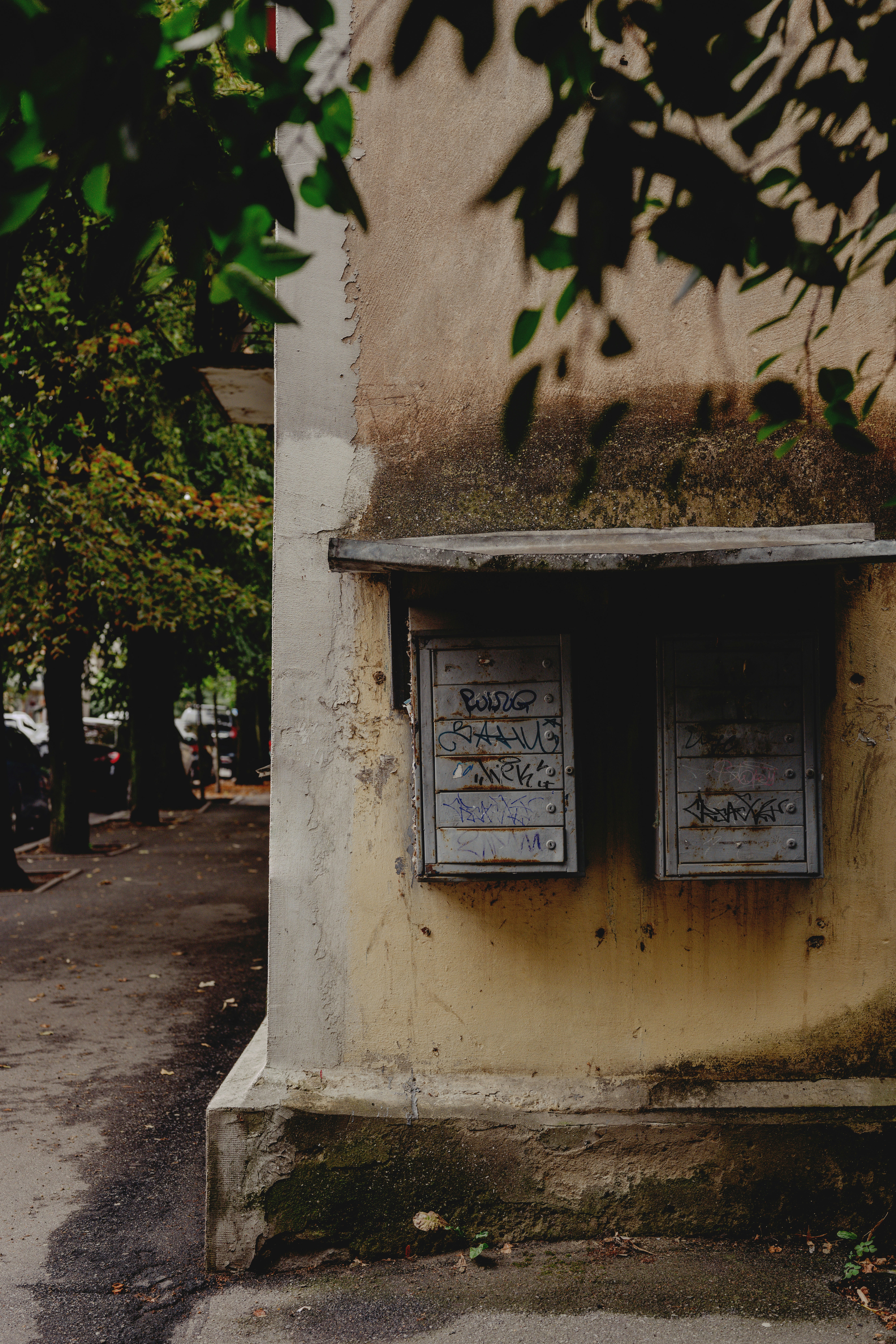 Two weathered metal mailboxes with graffiti on a beige building corner near trees