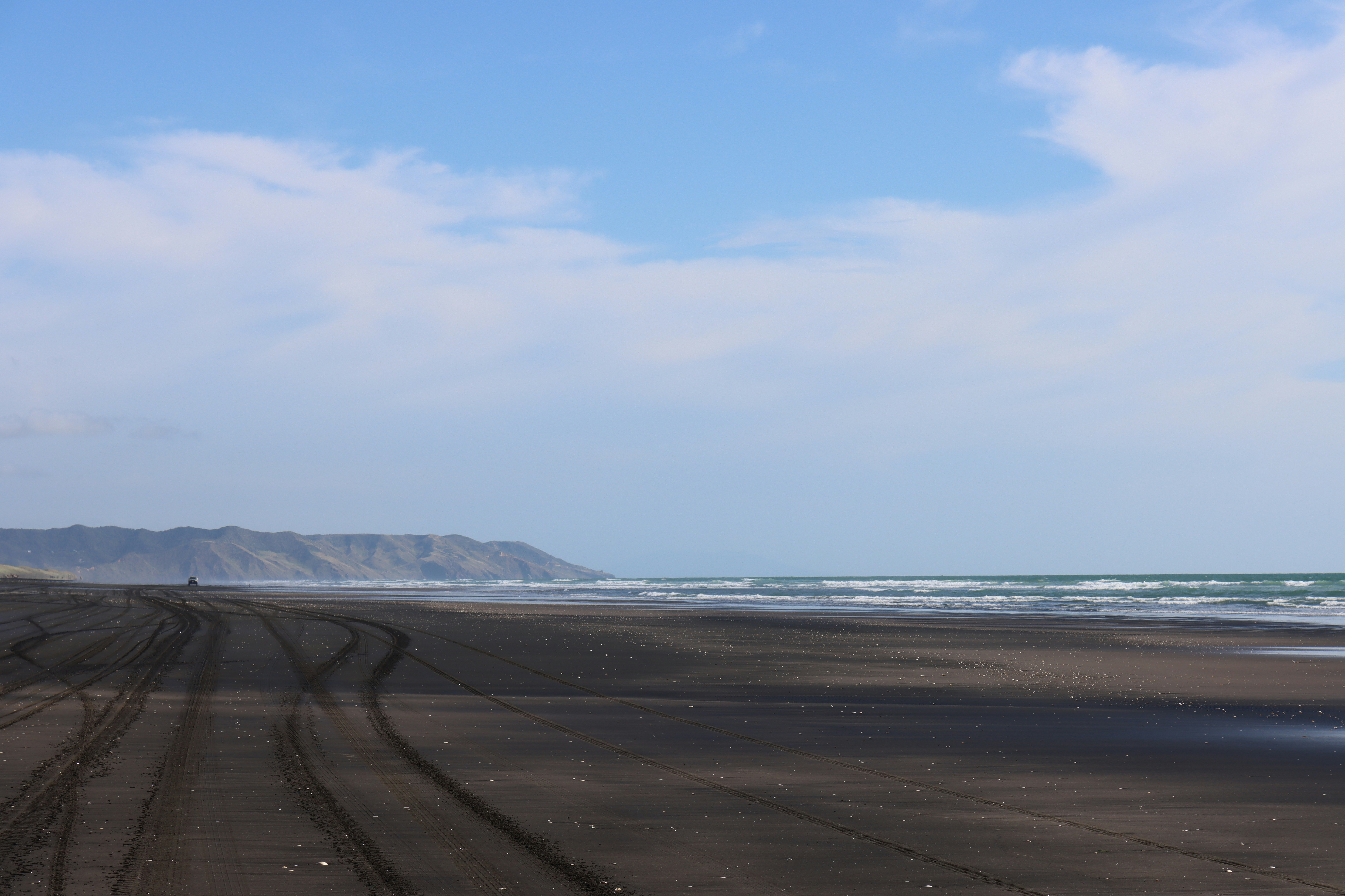 Tire tracks on a dark sand beach with distant hills and ocean waves
