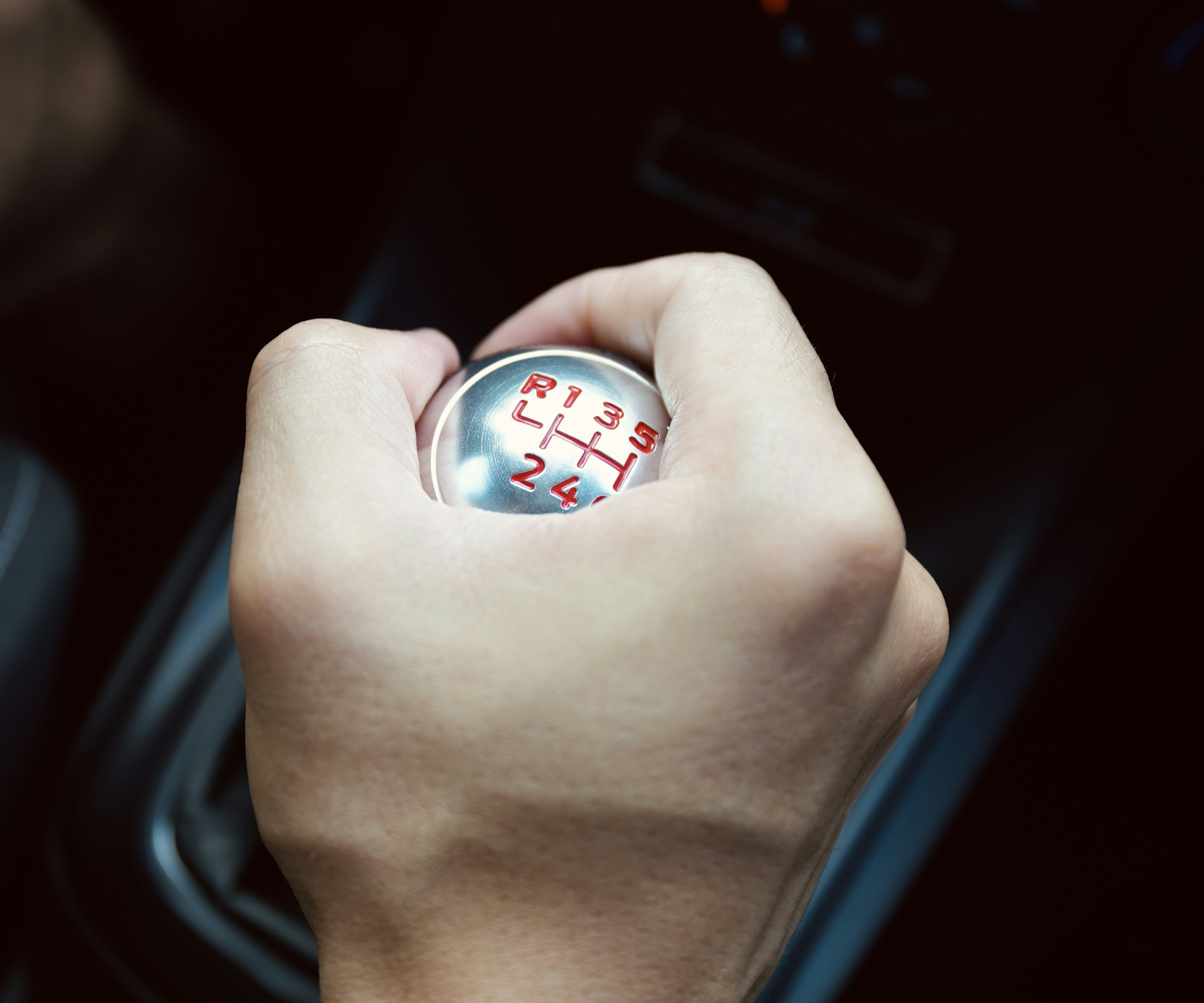A hand gripping a metallic manual gear shift knob with red gear markings