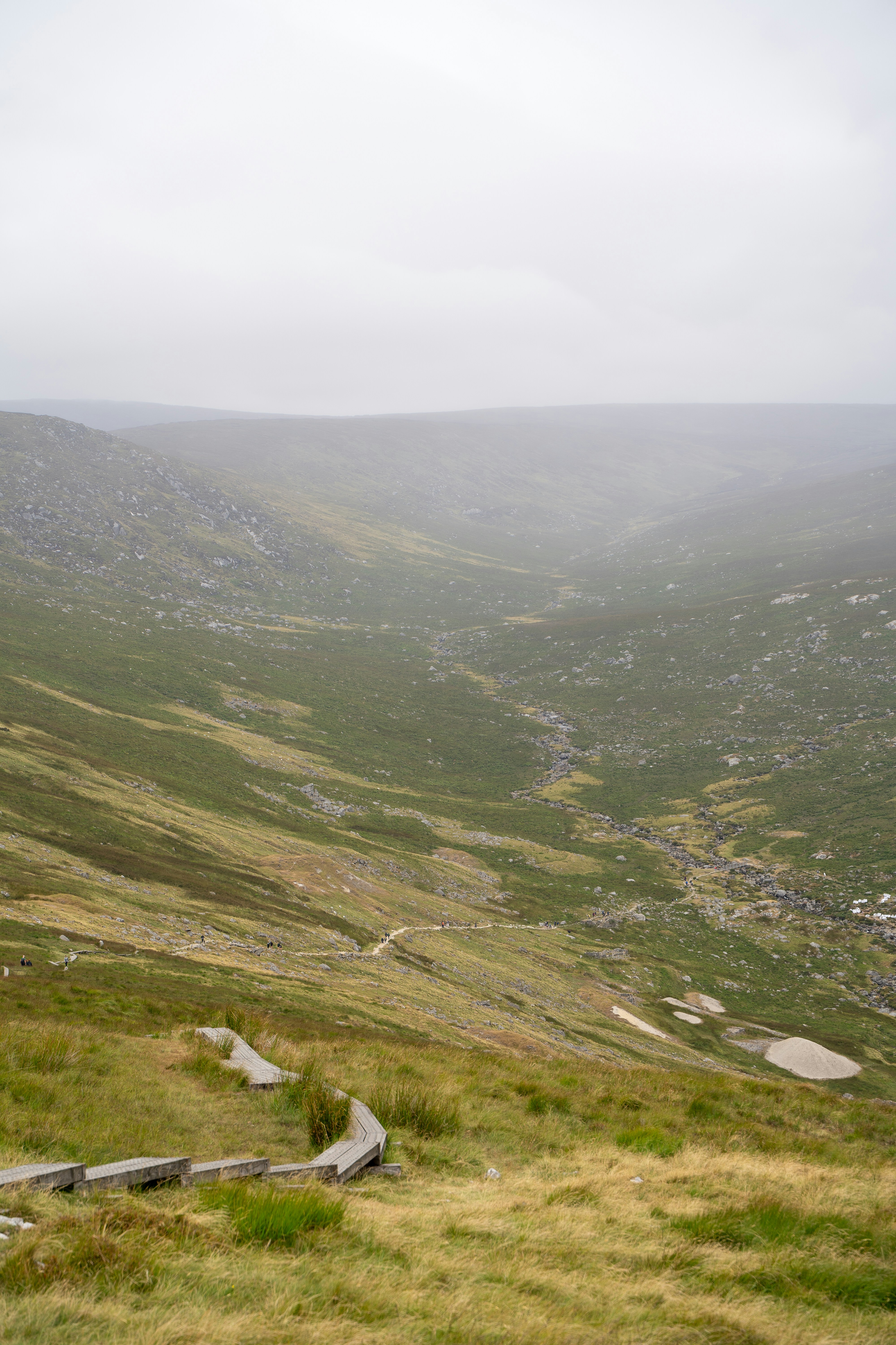 A wooden boardwalk winding through a misty green valley with rocky slopes