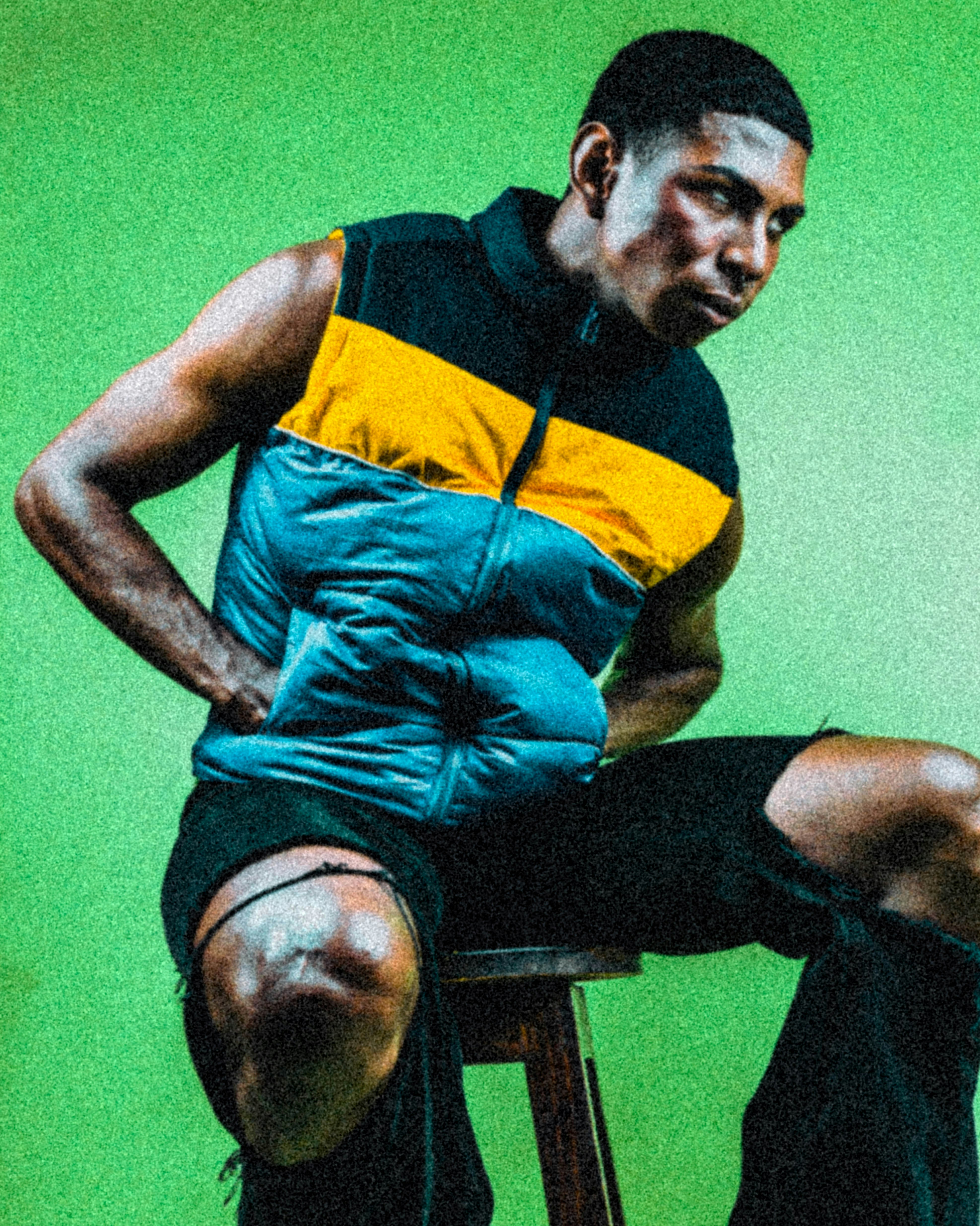 A man in a color-blocked vest sitting on a stool against a green background
