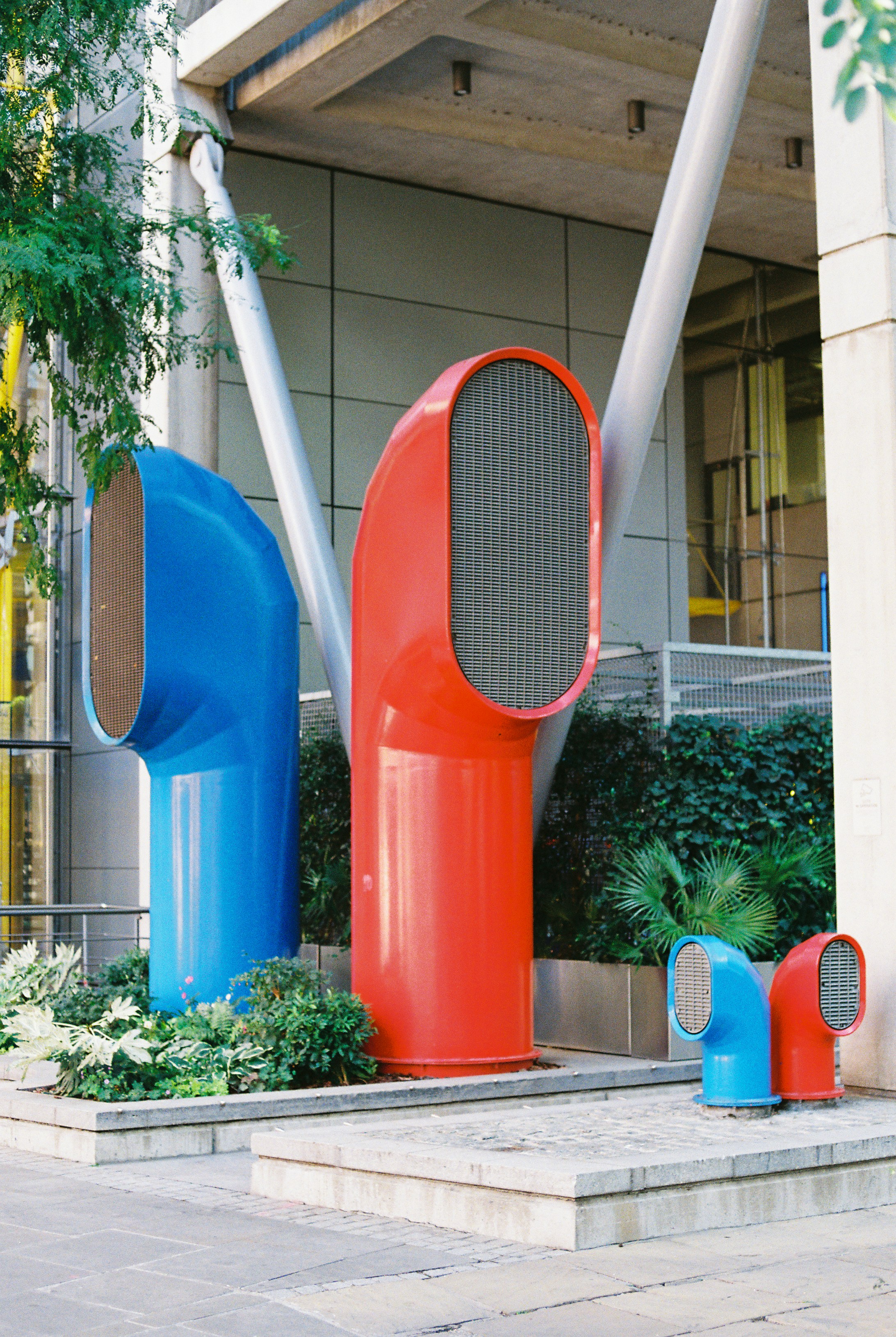 Large blue and red ventilation ducts standing among green plants outside a building