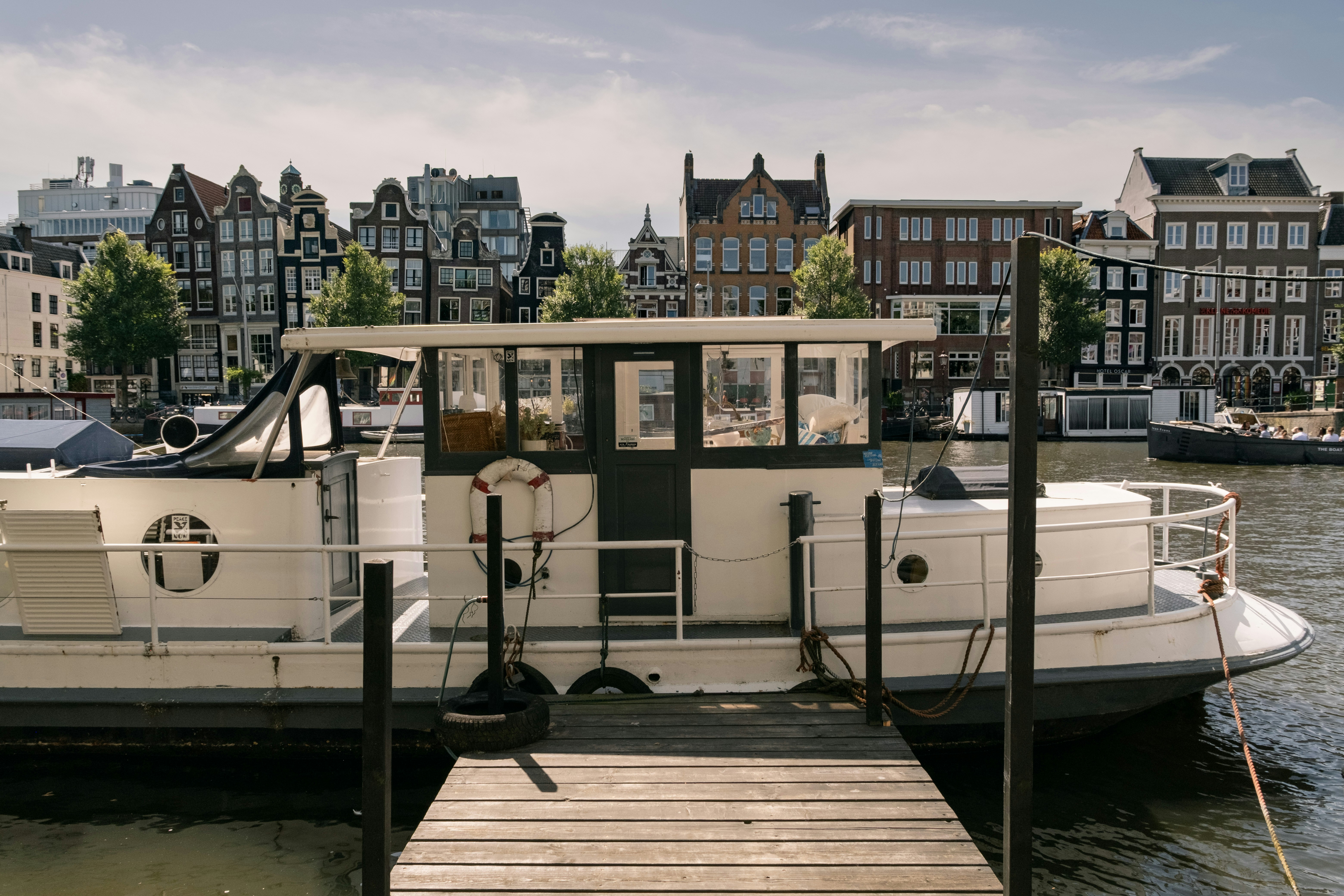 A white houseboat moored at a wooden dock in Amsterdam with historic canal houses