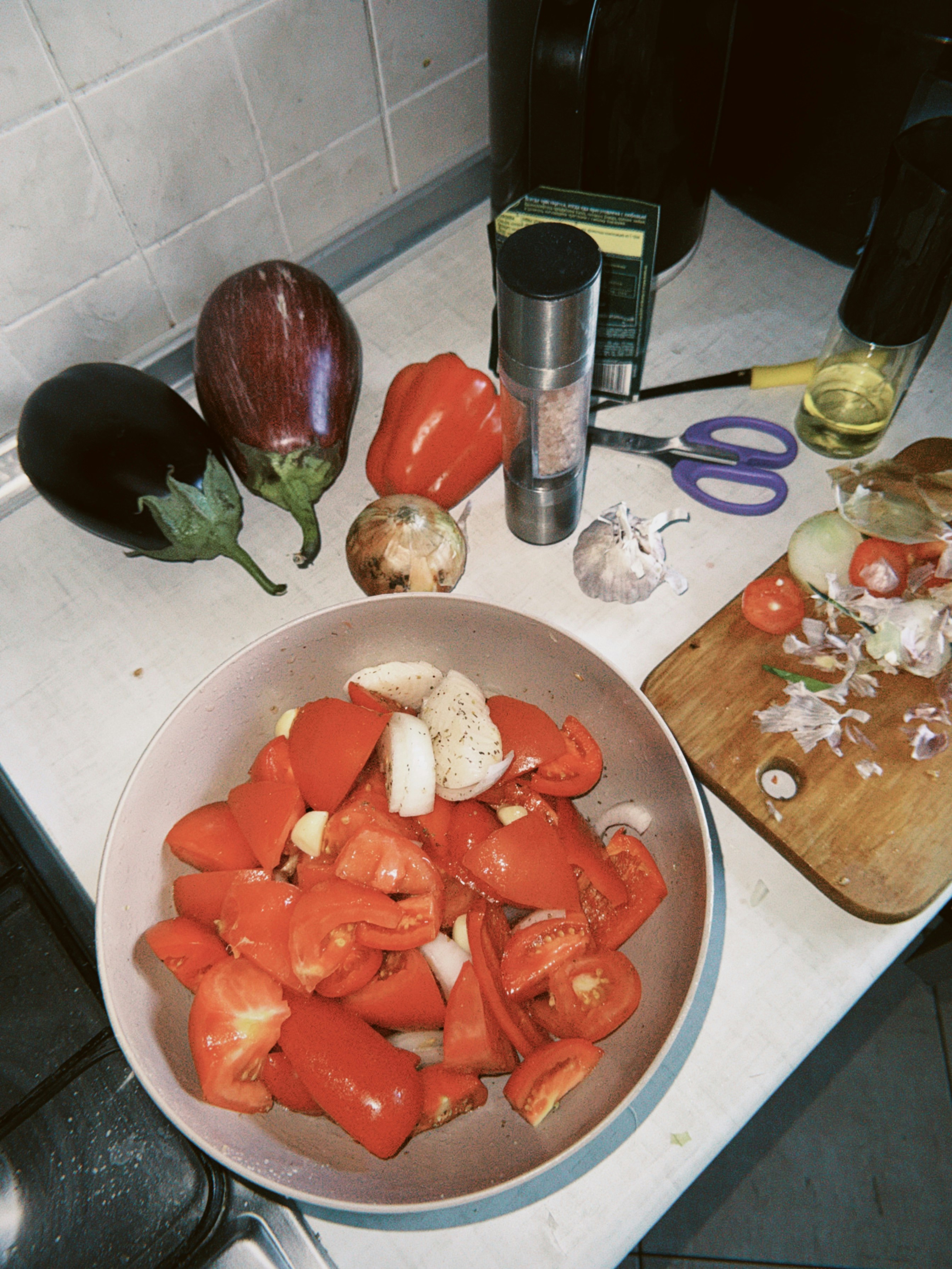 A bowl of chopped tomatoes and onions beside fresh eggplants, bell pepper, and garlic