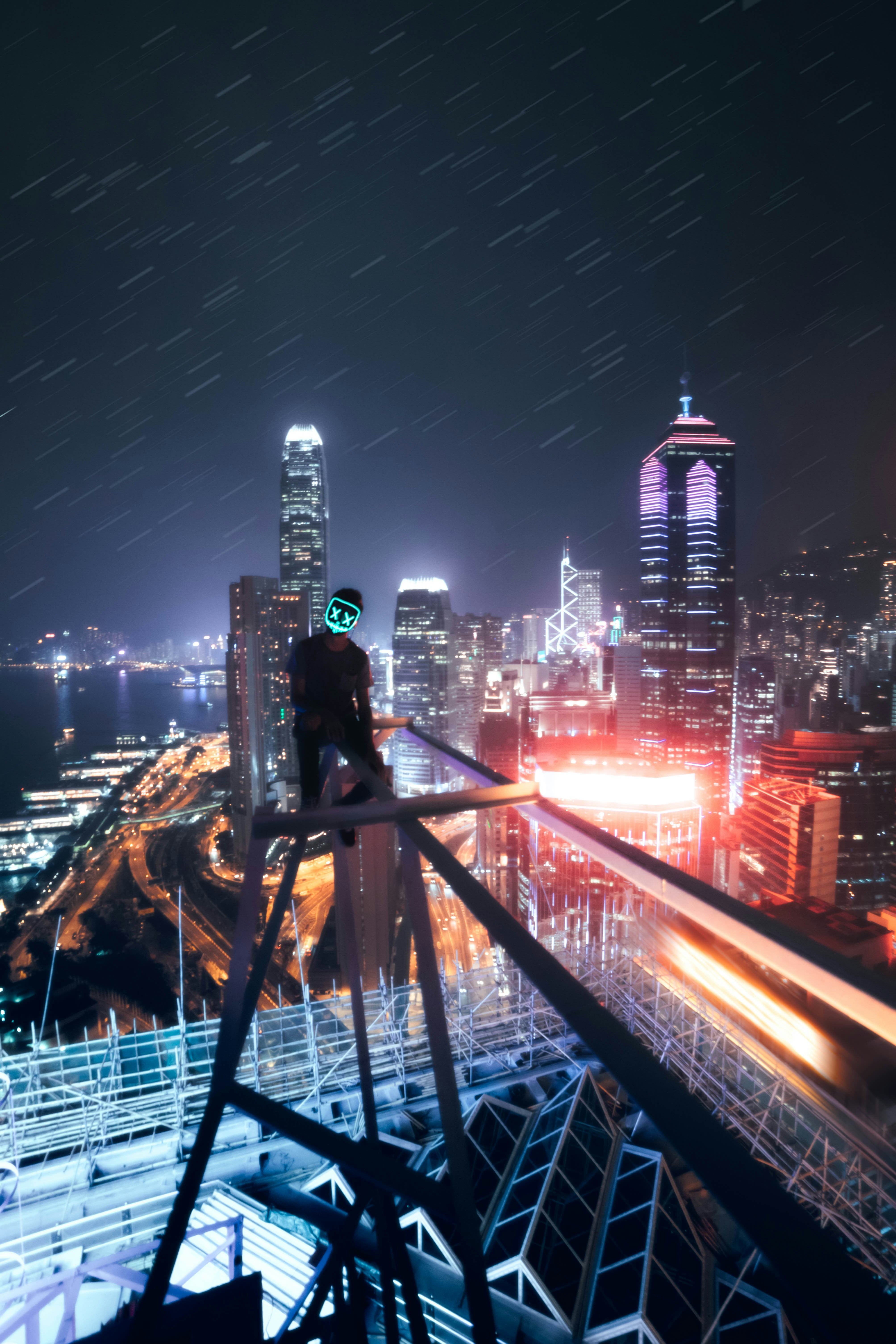 A person wearing a glowing neon mask on a skyscraper overlooking Hong Kong