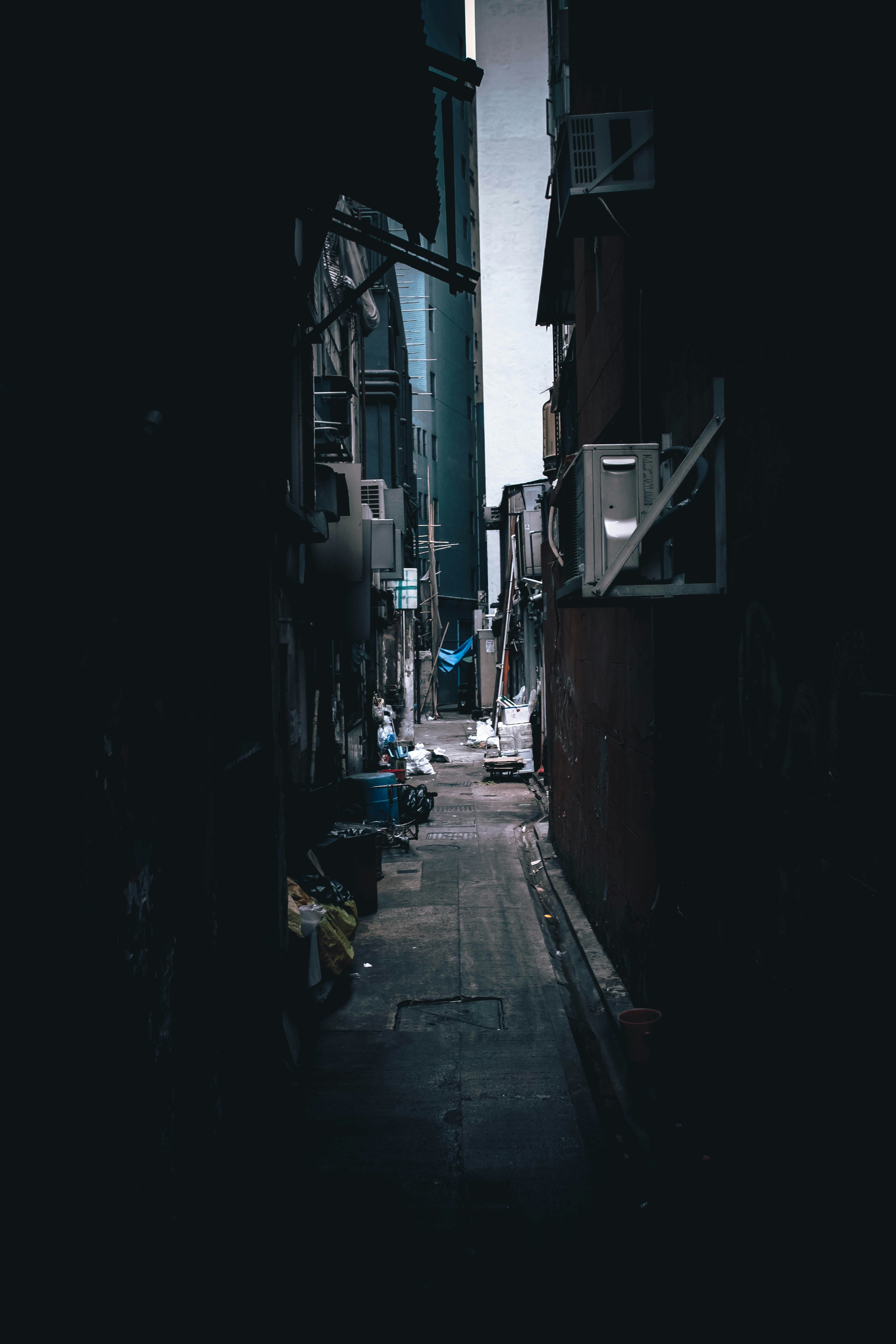A narrow urban alleyway between tall buildings with air conditioning units and debris