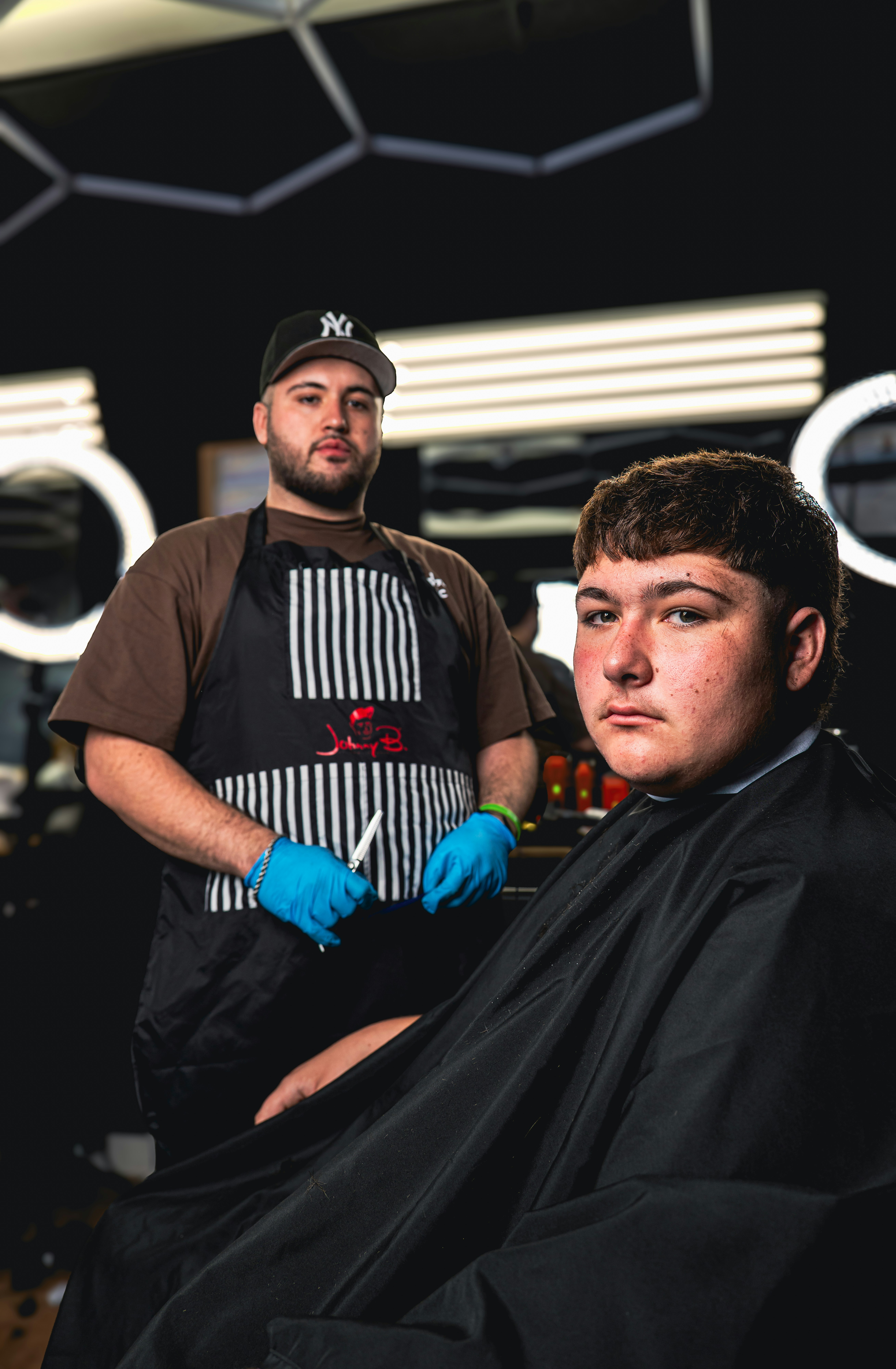 A barber in a striped apron and blue gloves standing behind a seated client
