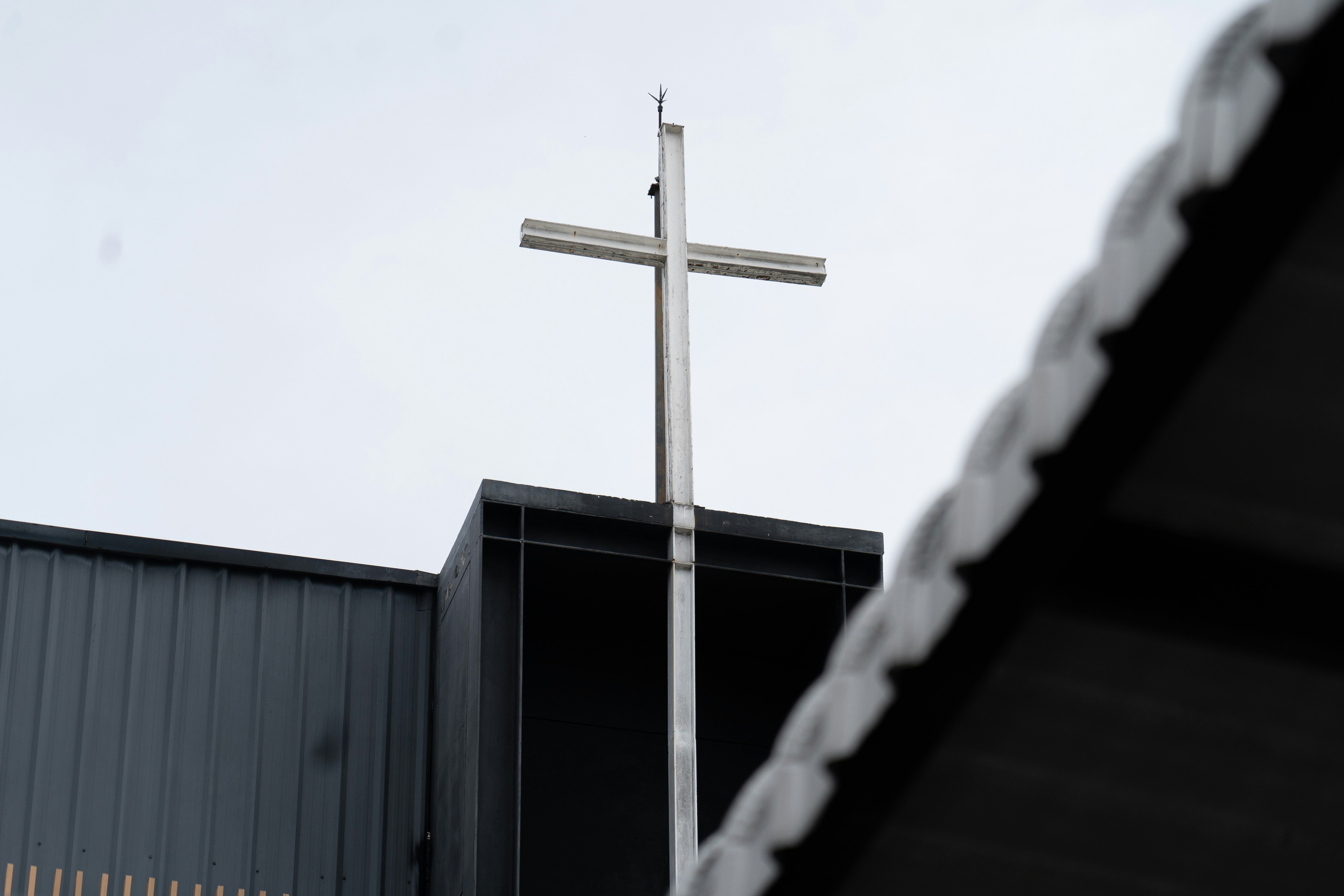 A white metal cross atop a modern dark building against a pale sky