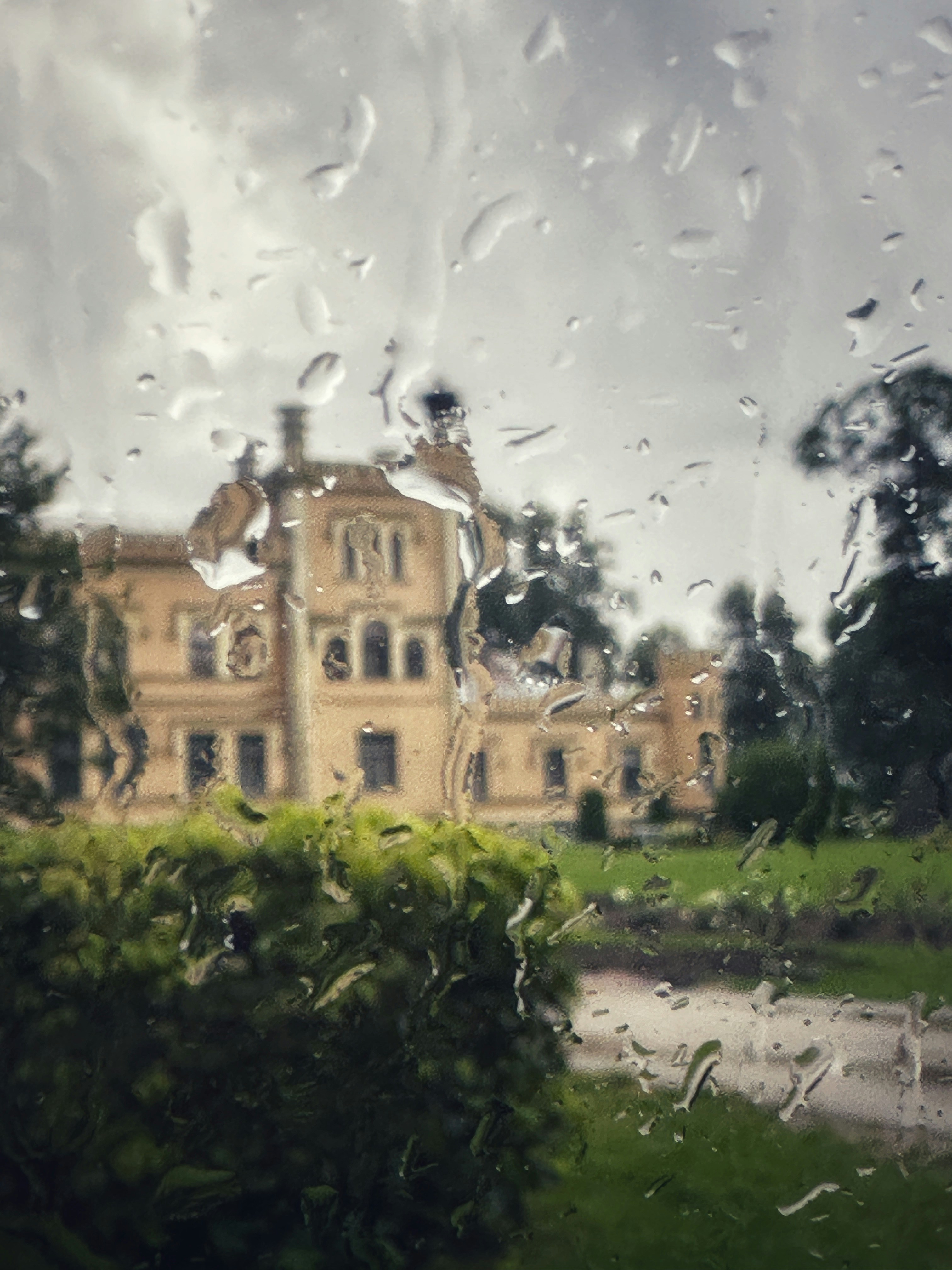 A historic stone mansion viewed through a rain-streaked window with green garden foliage