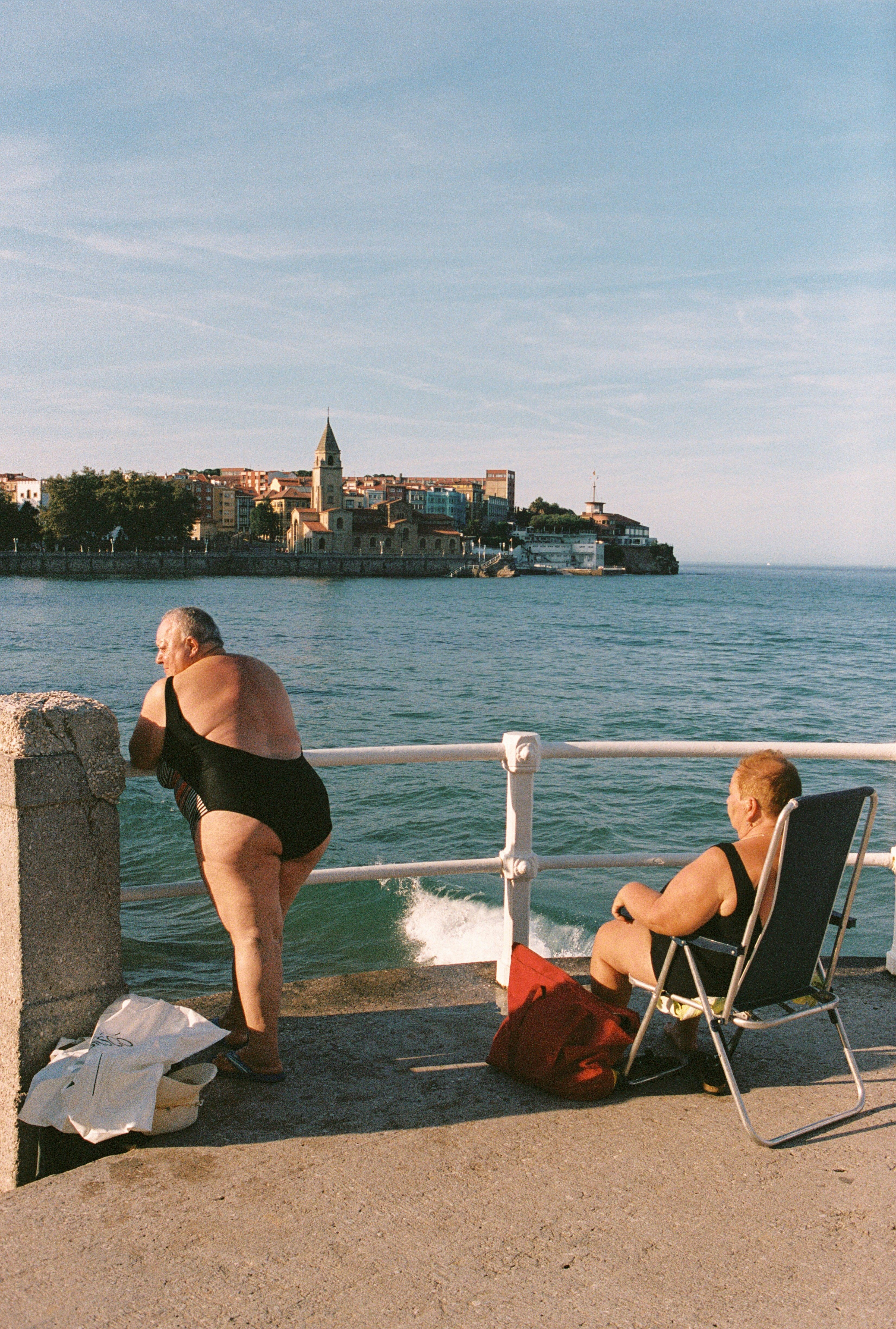 Two women in swimsuits on a pier overlooking the Gijón coastline in Spain