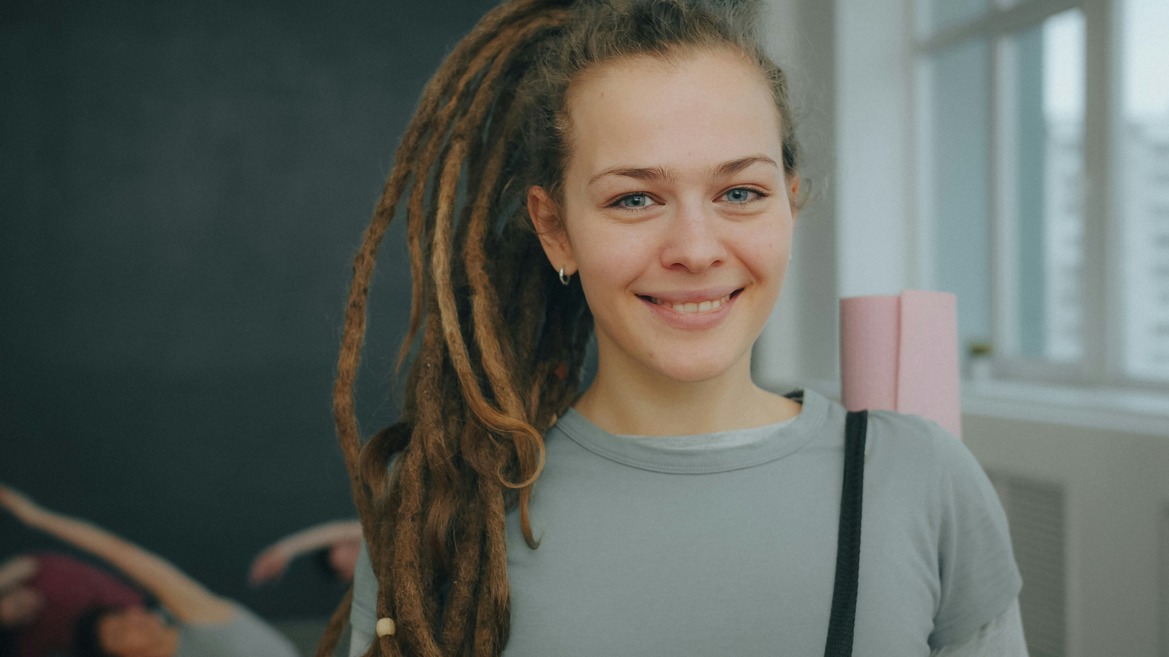 A smiling woman with long dreadlocks carrying a pink yoga mat in a studio