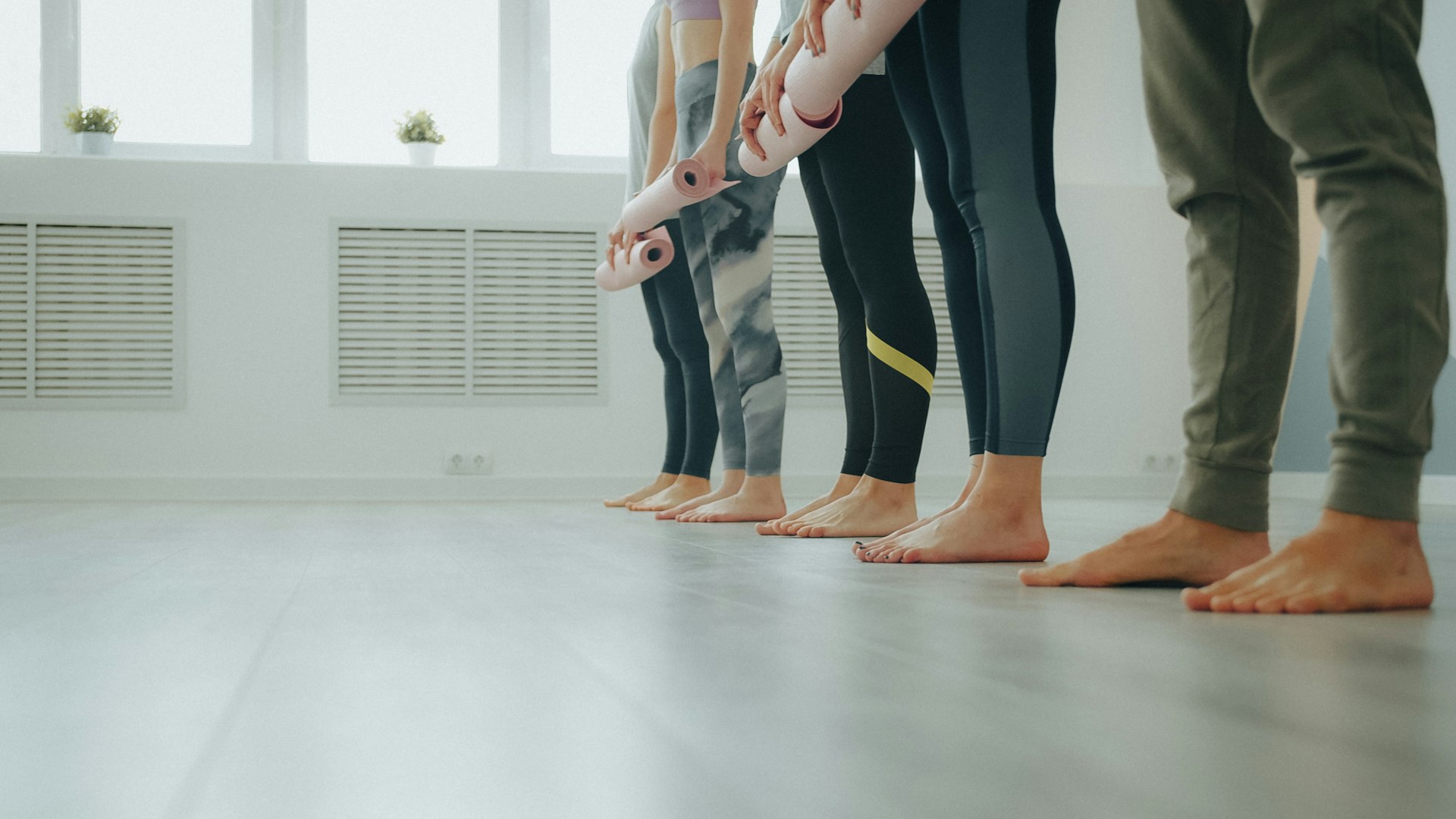 Students holding rolled yoga mats as they line up barefoot in a bright studio