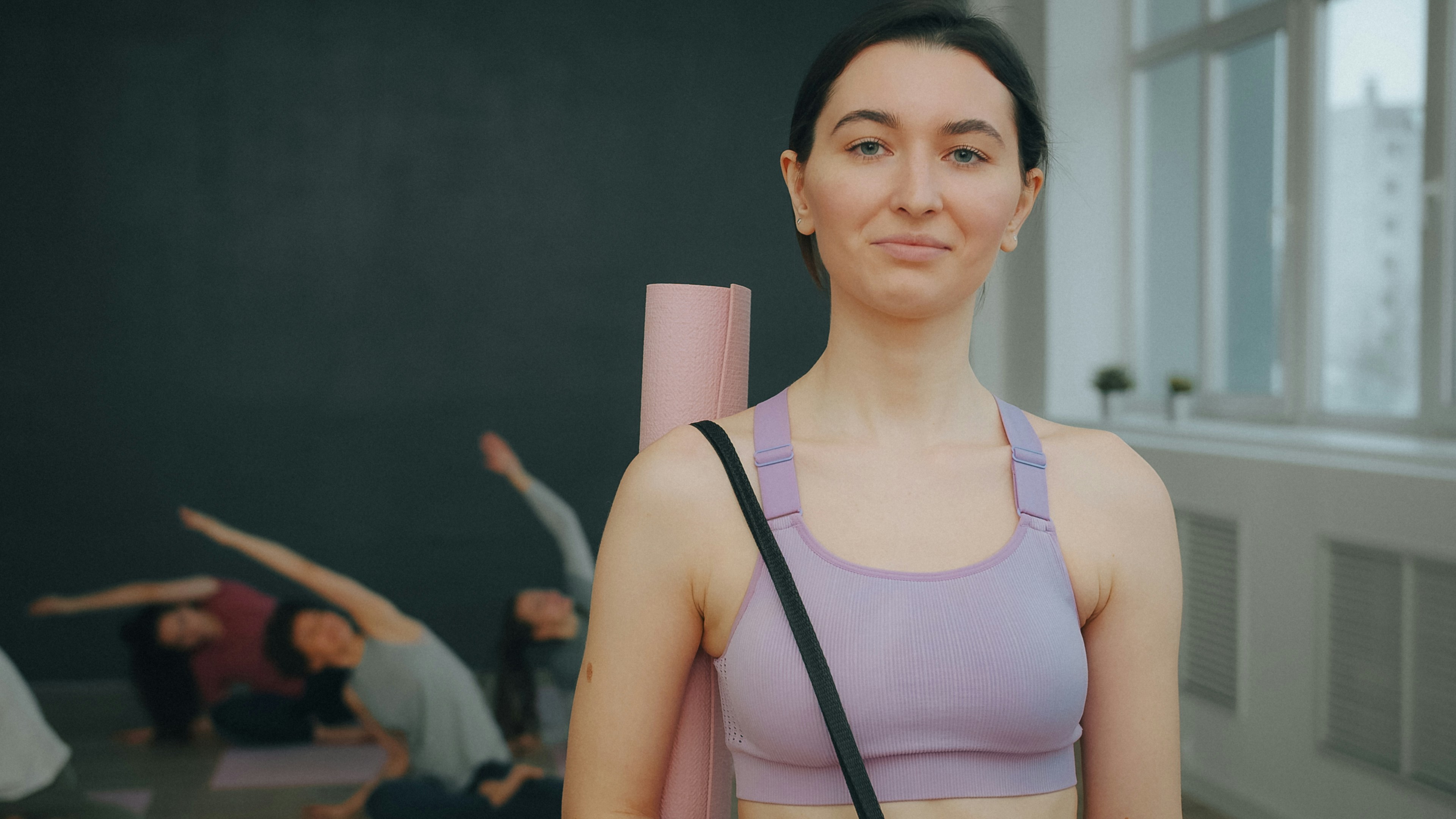 A woman in a lavender sports bra holding a pink yoga mat in a studio