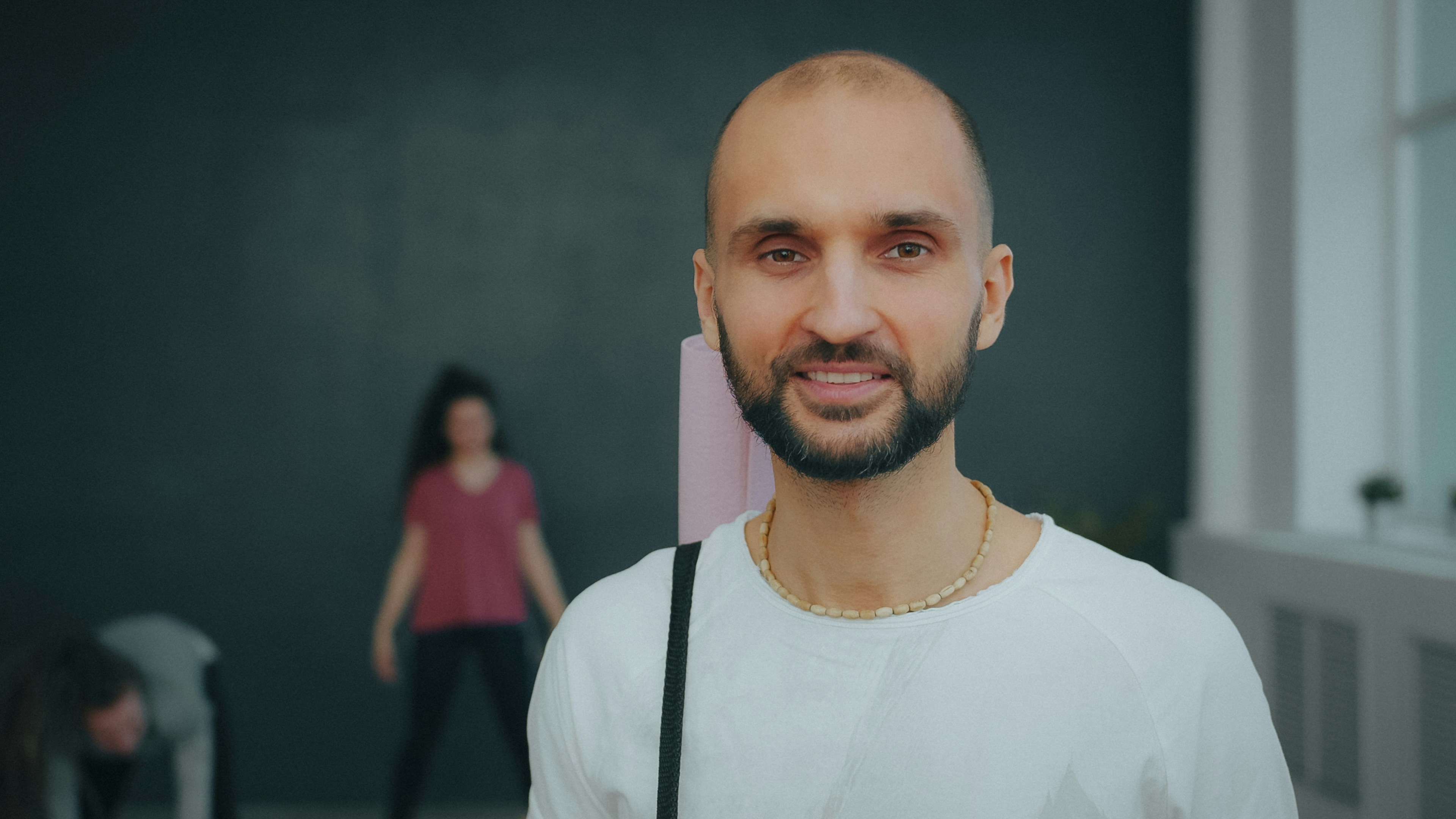 A smiling man with a beard carrying a pink yoga mat in a studio