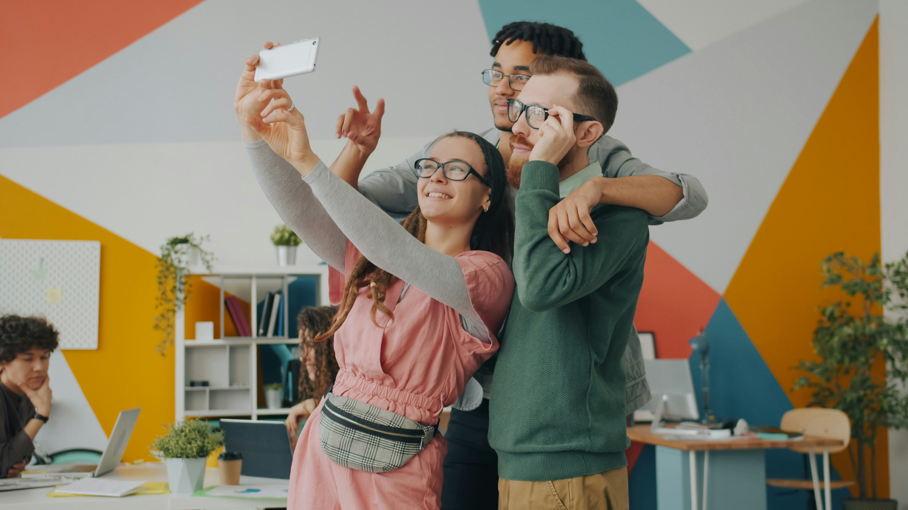 Happy young people girl and guys are taking selfie at work together using modern smartphone camera posing with v-sign and thumbs-up hand gestures