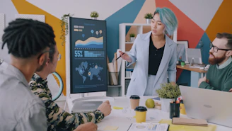 A woman with blue hair presenting data on a monitor to a team