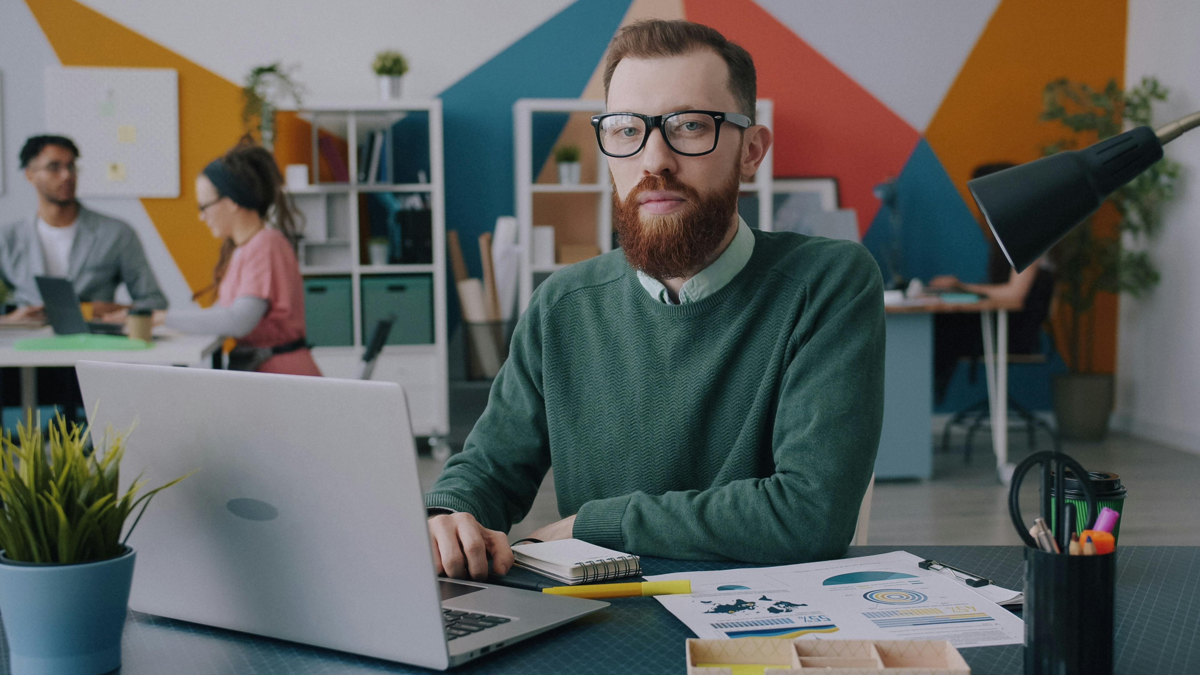 A man with a beard and glasses working at a laptop in an office