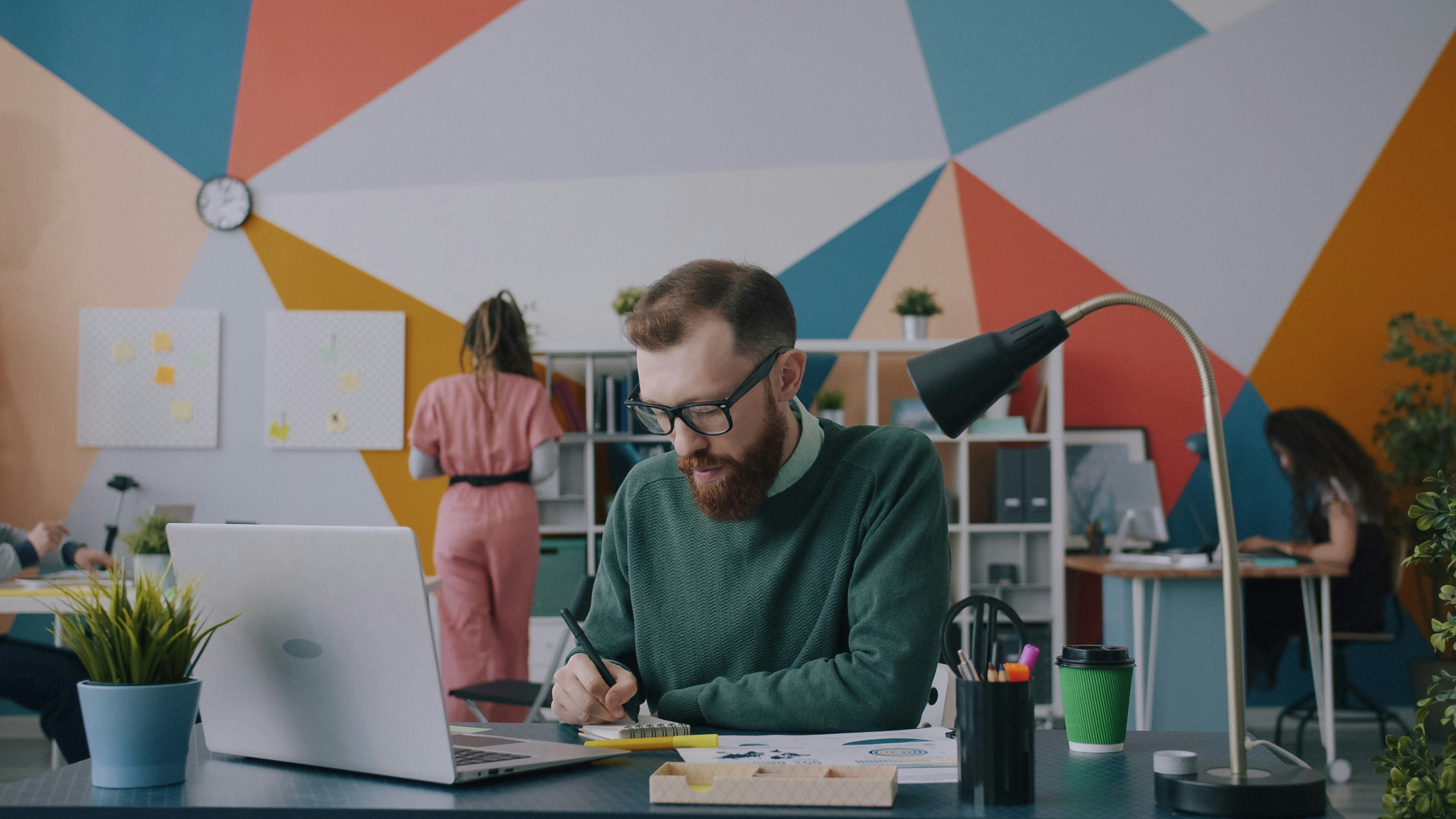 Serious guy is using laptop computer and writing notes working in open space office with colleagues. Modern business and creative youth concept.
