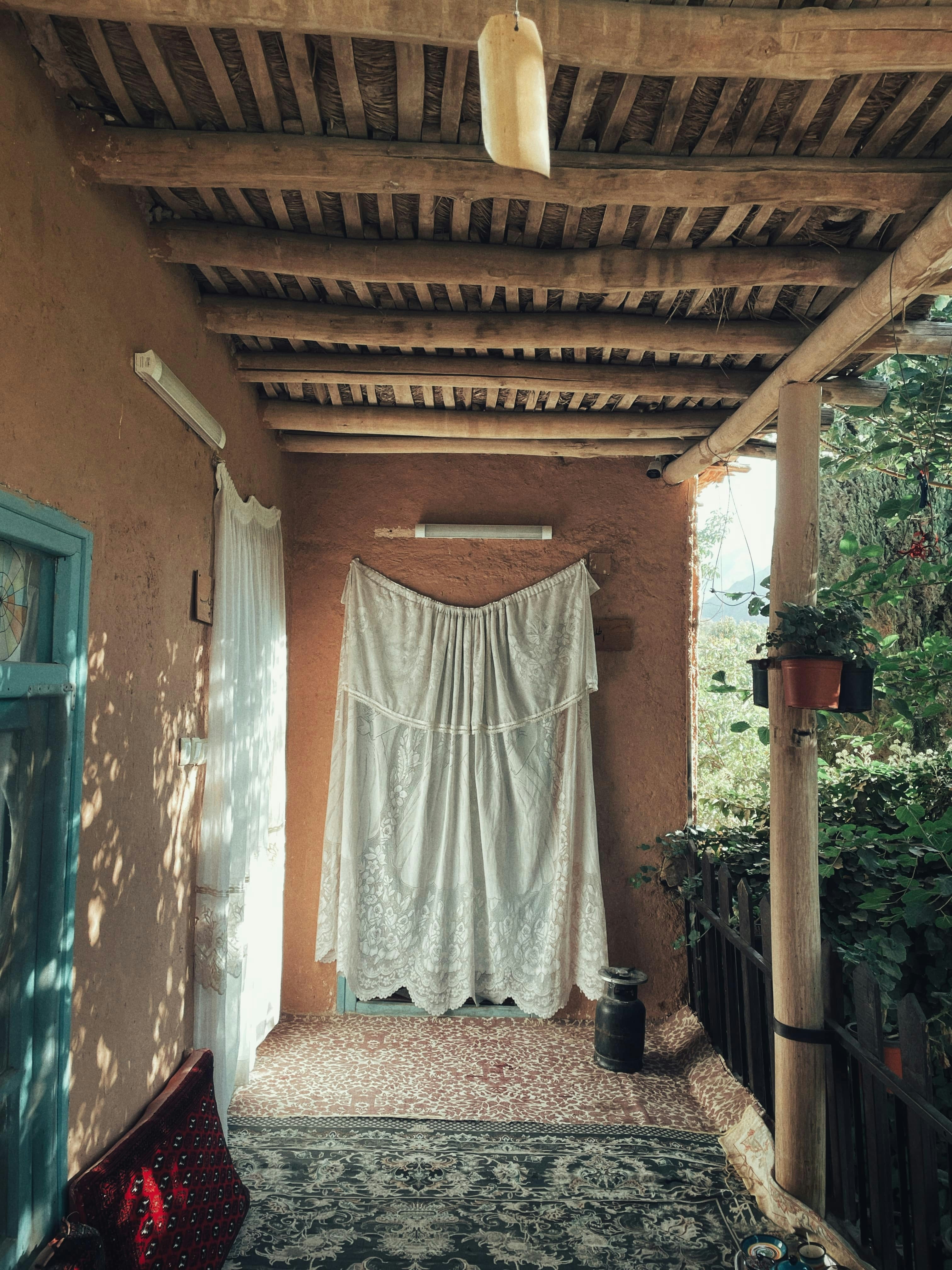 A rustic porch with mud walls, a thatched roof, and white lace curtains