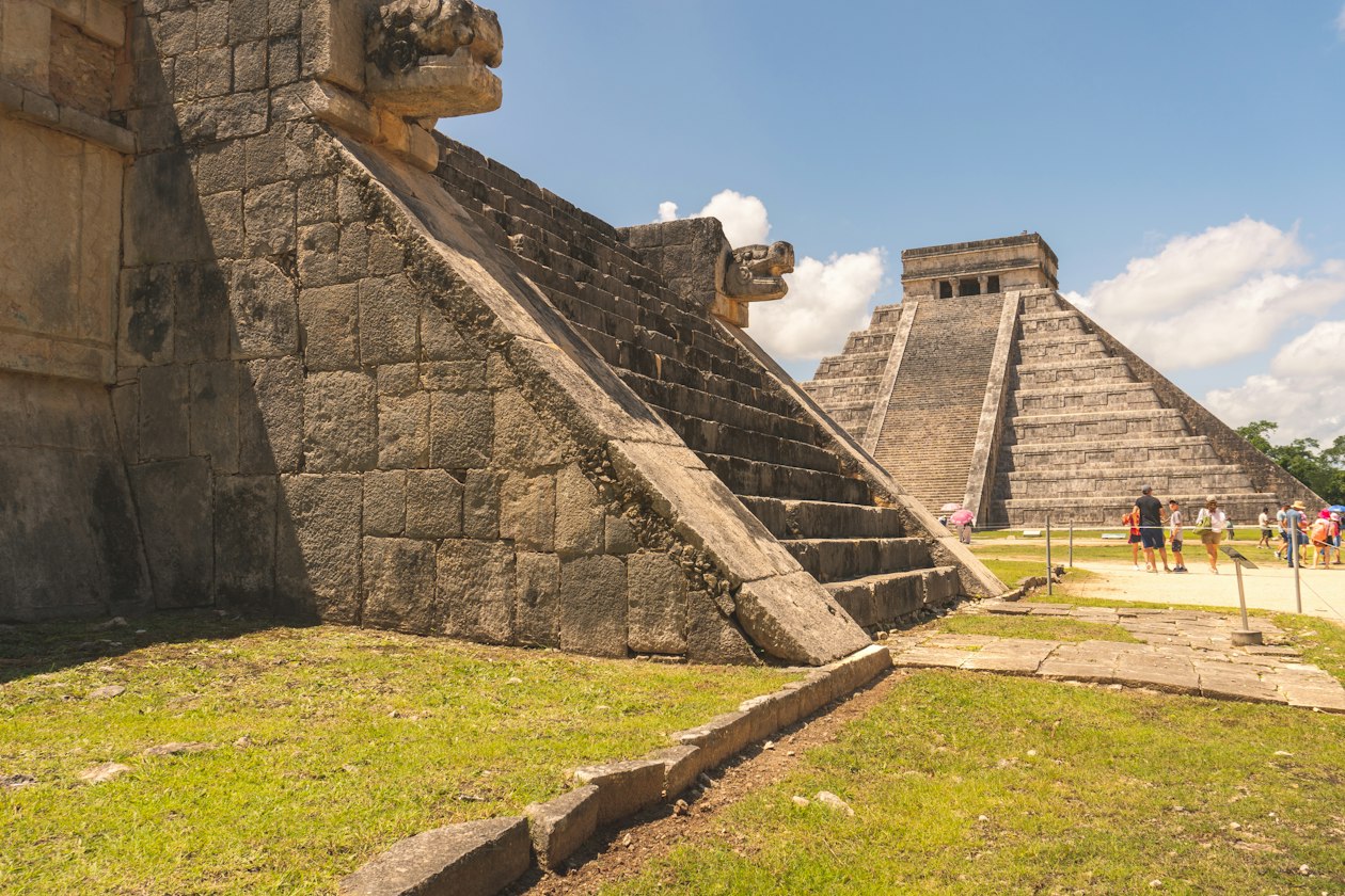 Stone serpent heads at Chichen Itza with El Castillo behind, the pyramid where the equinox serpent descends