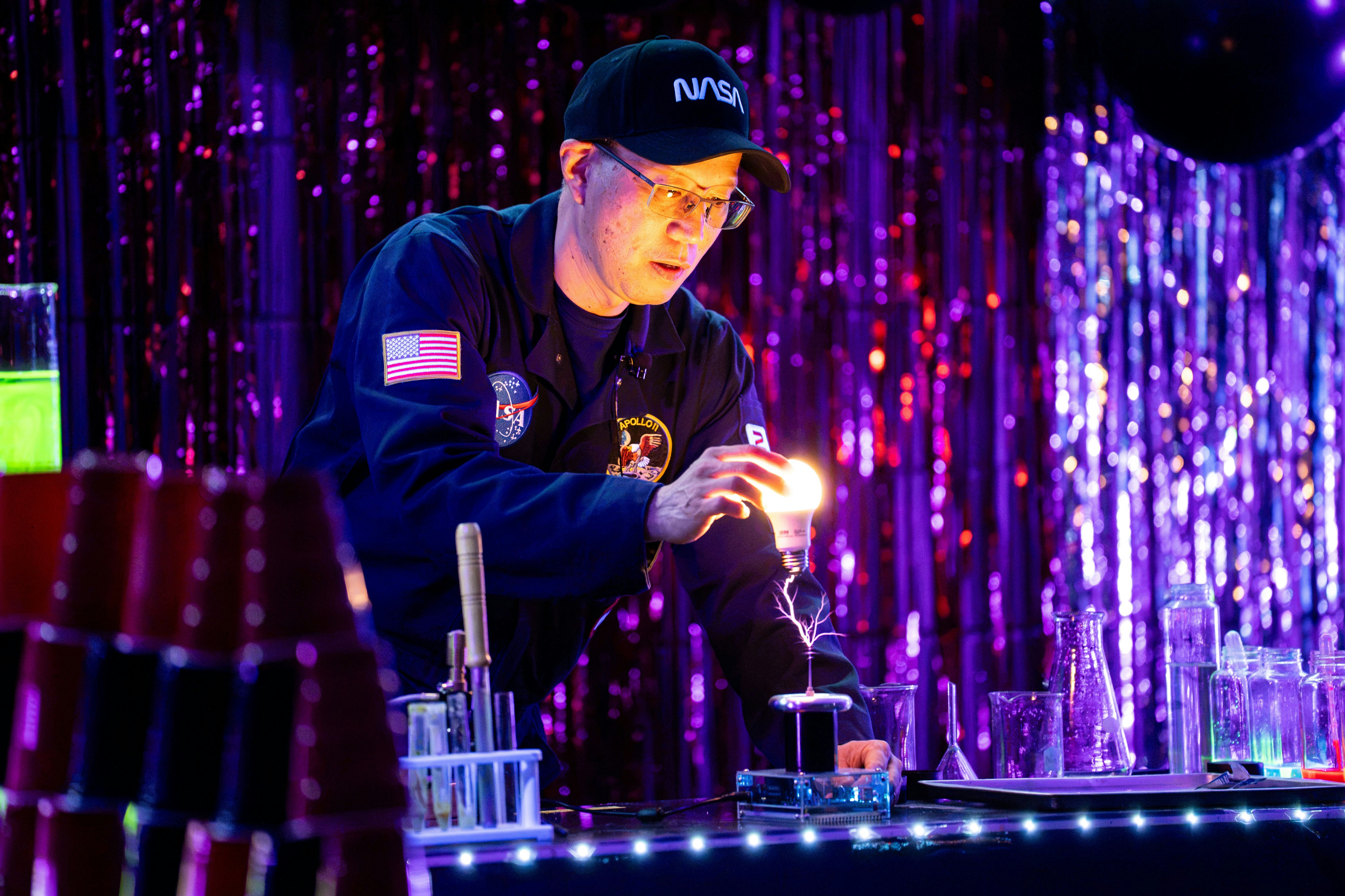 A person in a NASA flight suit holding a glowing lightbulb above a plasma ball