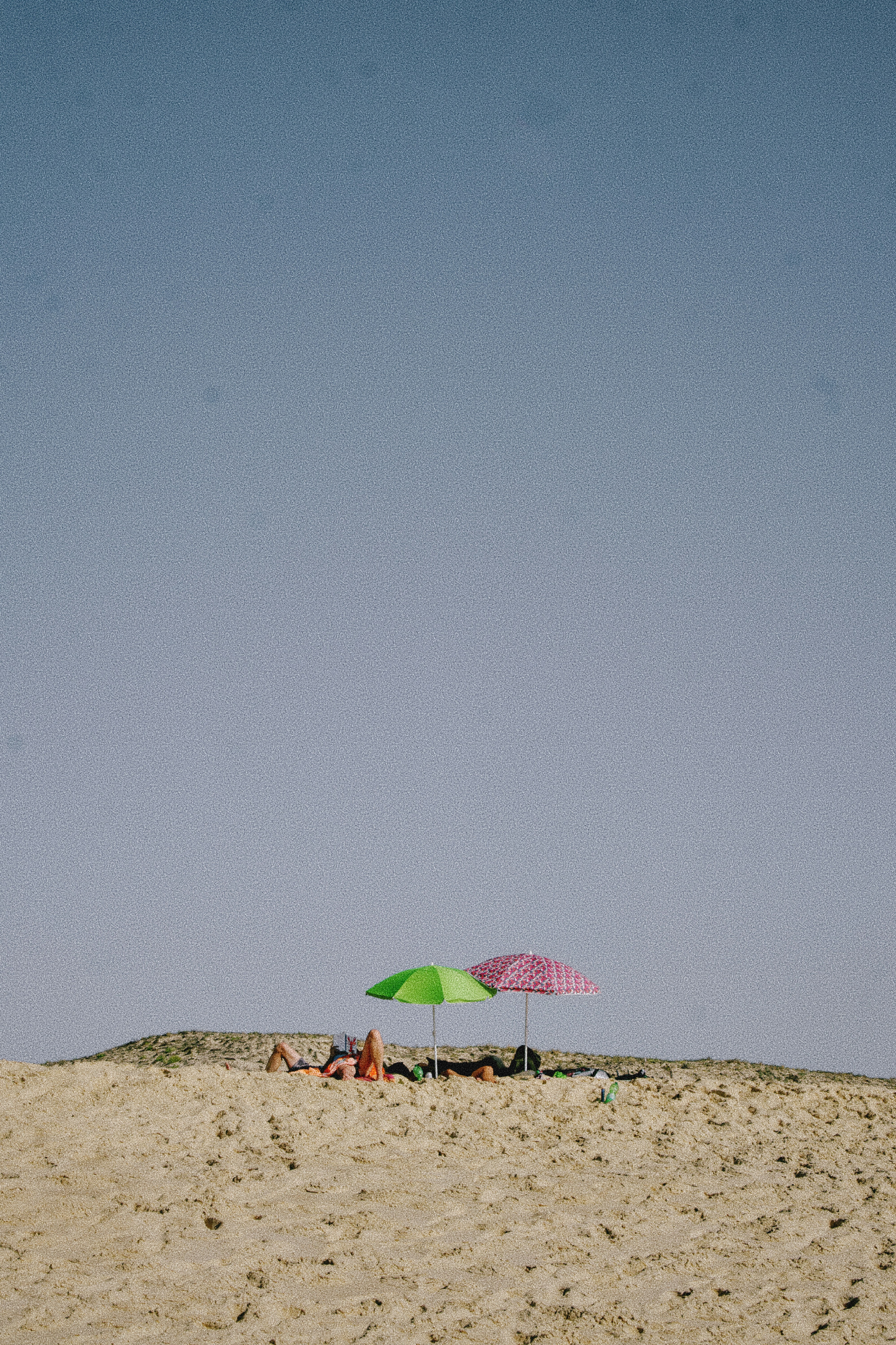 People relaxing under a green and a patterned pink umbrella on a sandy dune