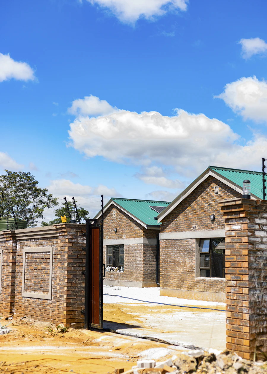 Brick houses with green metal roofs behind a boundary wall topped with electric fencing, beside an open steel gate.
