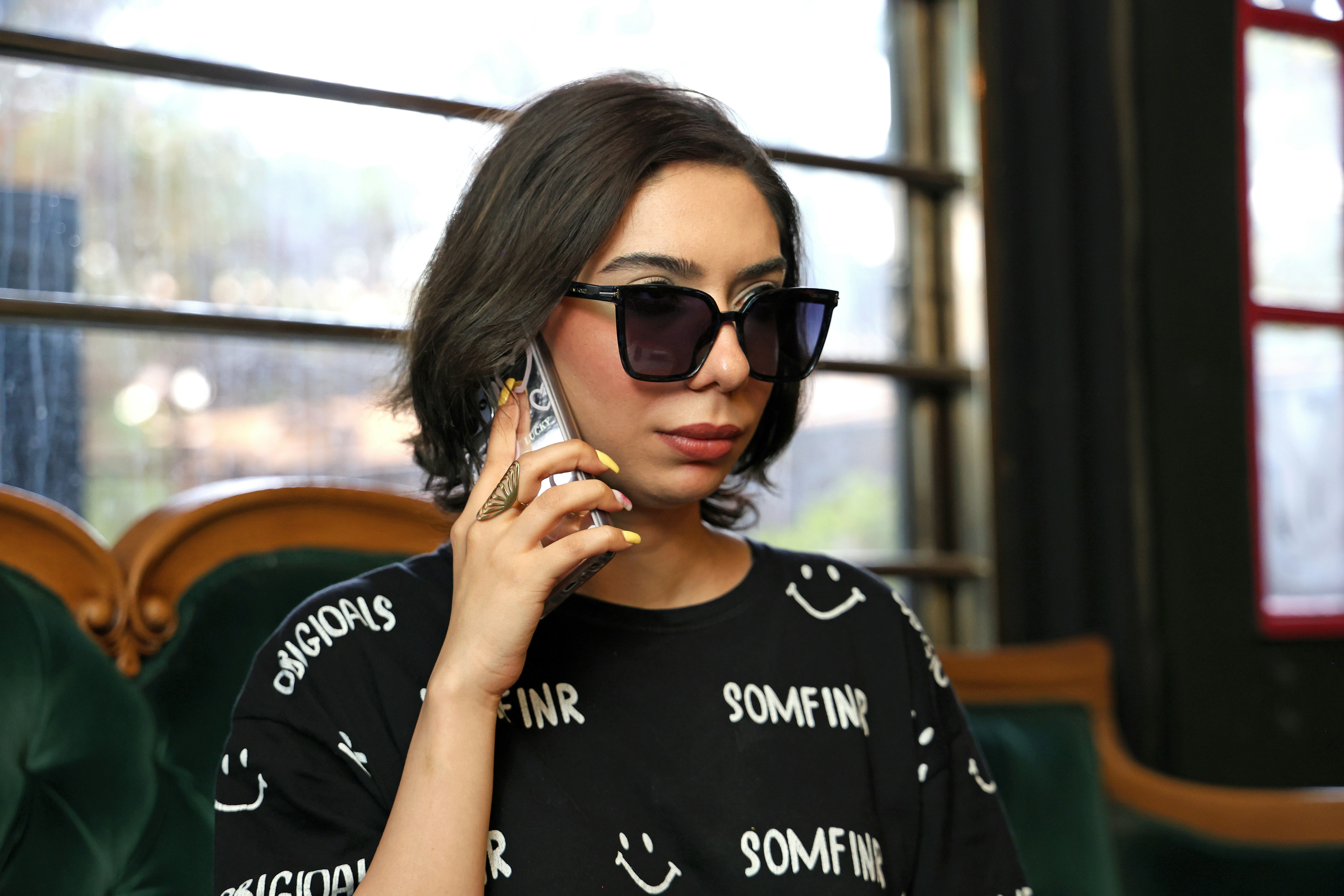 Una mujer de pelo oscuro con gafas de sol hablando por un smartphone en una cafetería