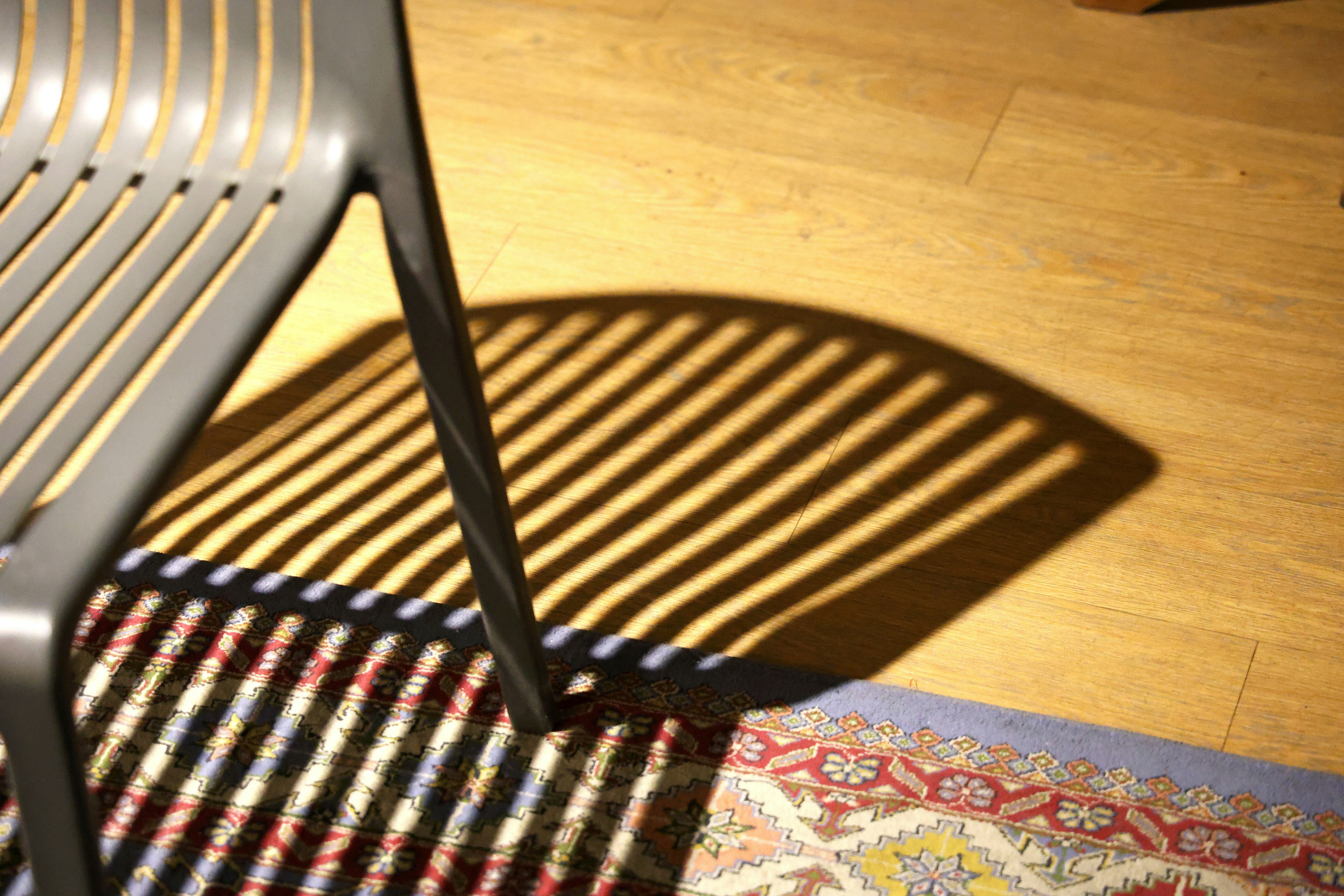 Una silla gris de listones que proyectaba largas sombras rayadas sobre una alfombra y el suelo con patrones