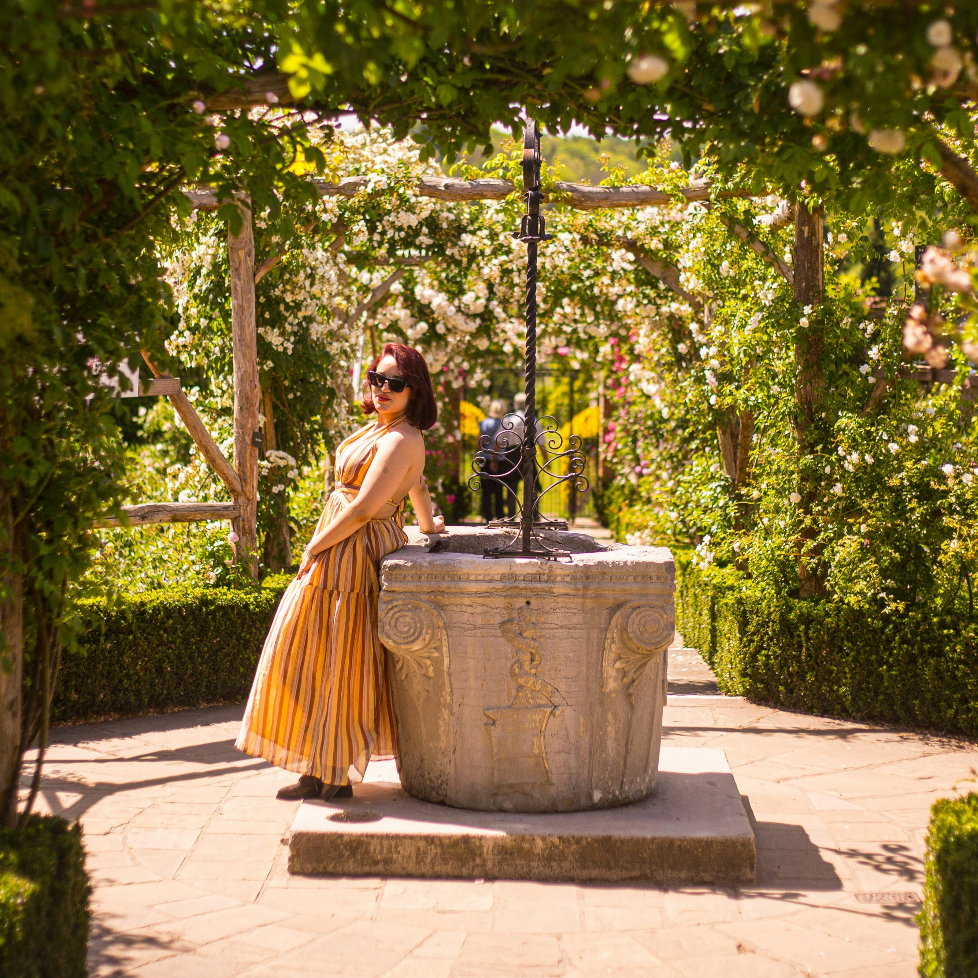 A woman in a striped dress leaning against a stone well in a rose garden