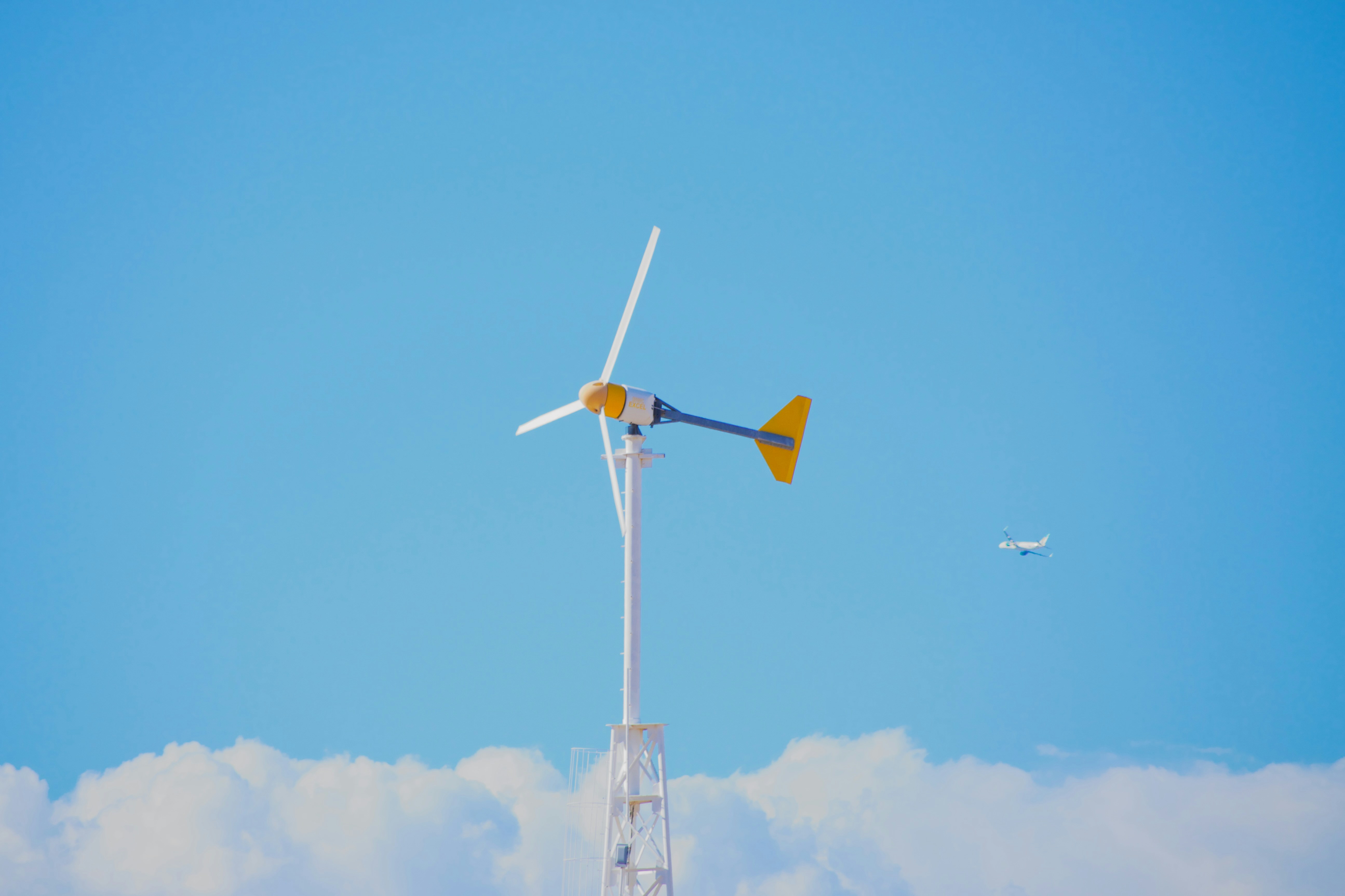 A small wind turbine against a clear blue sky with a distant airplane