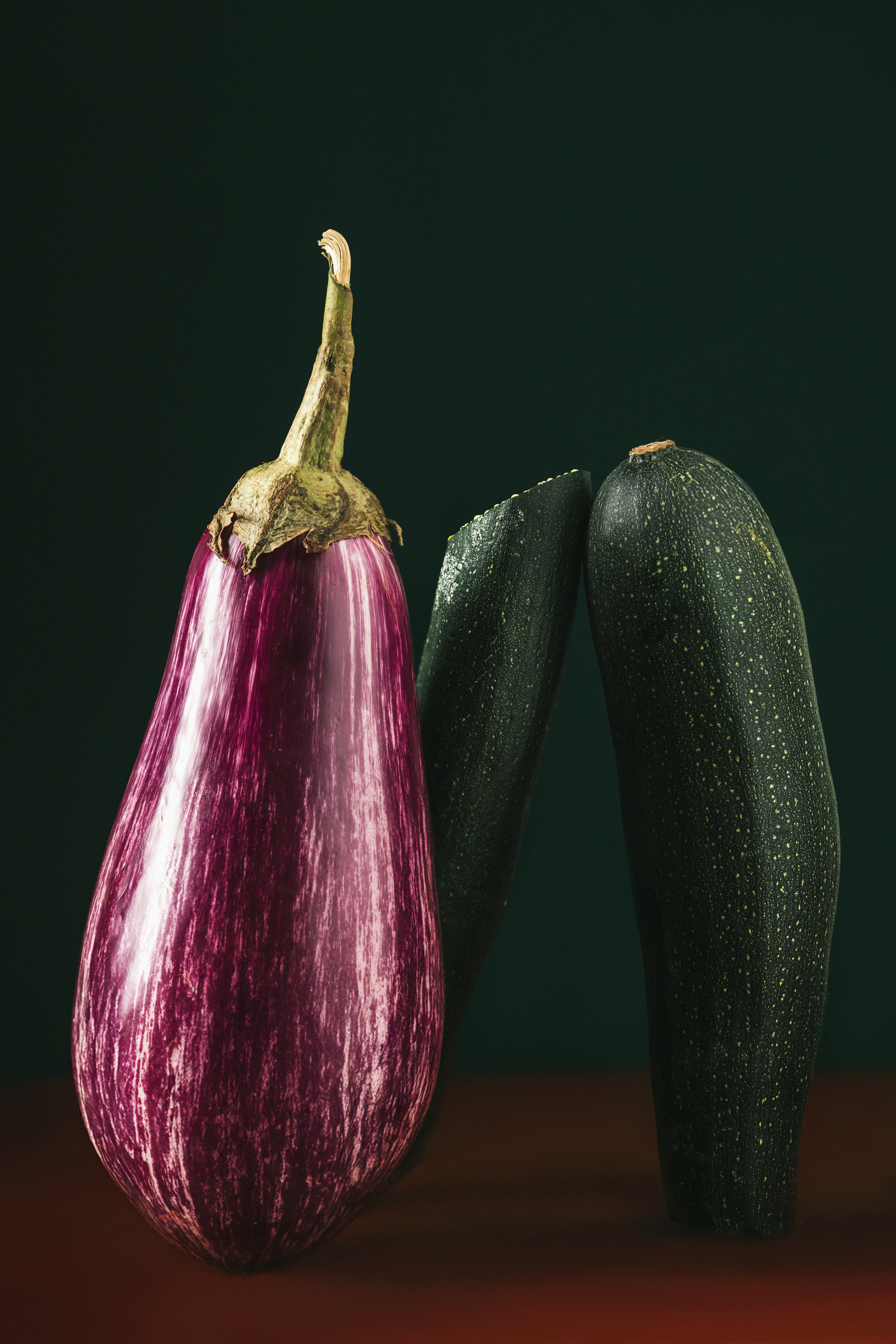 A striped purple eggplant beside a dark green zucchini on a dark surface