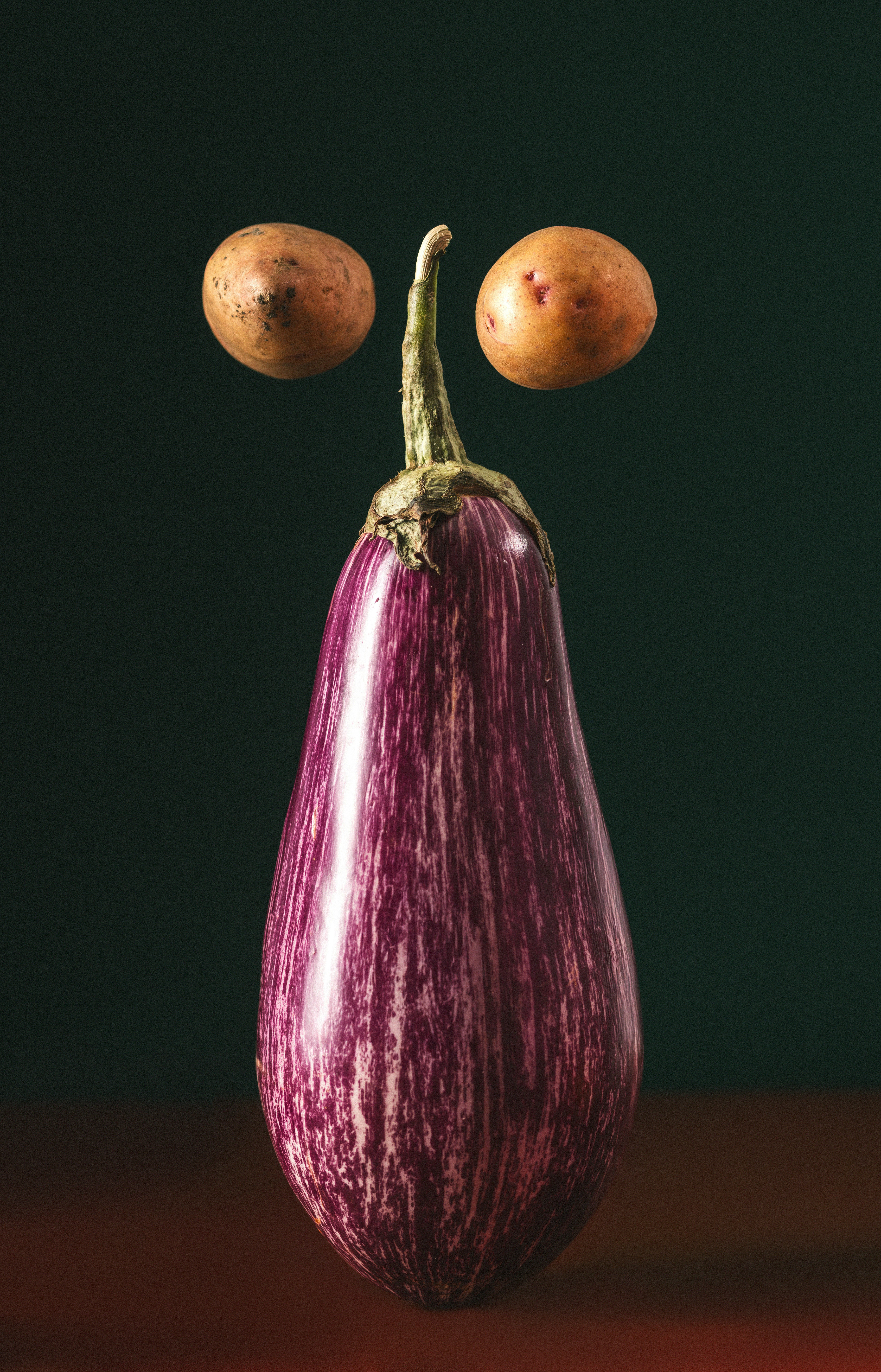 A striped purple eggplant with two small potatoes above it on a dark background