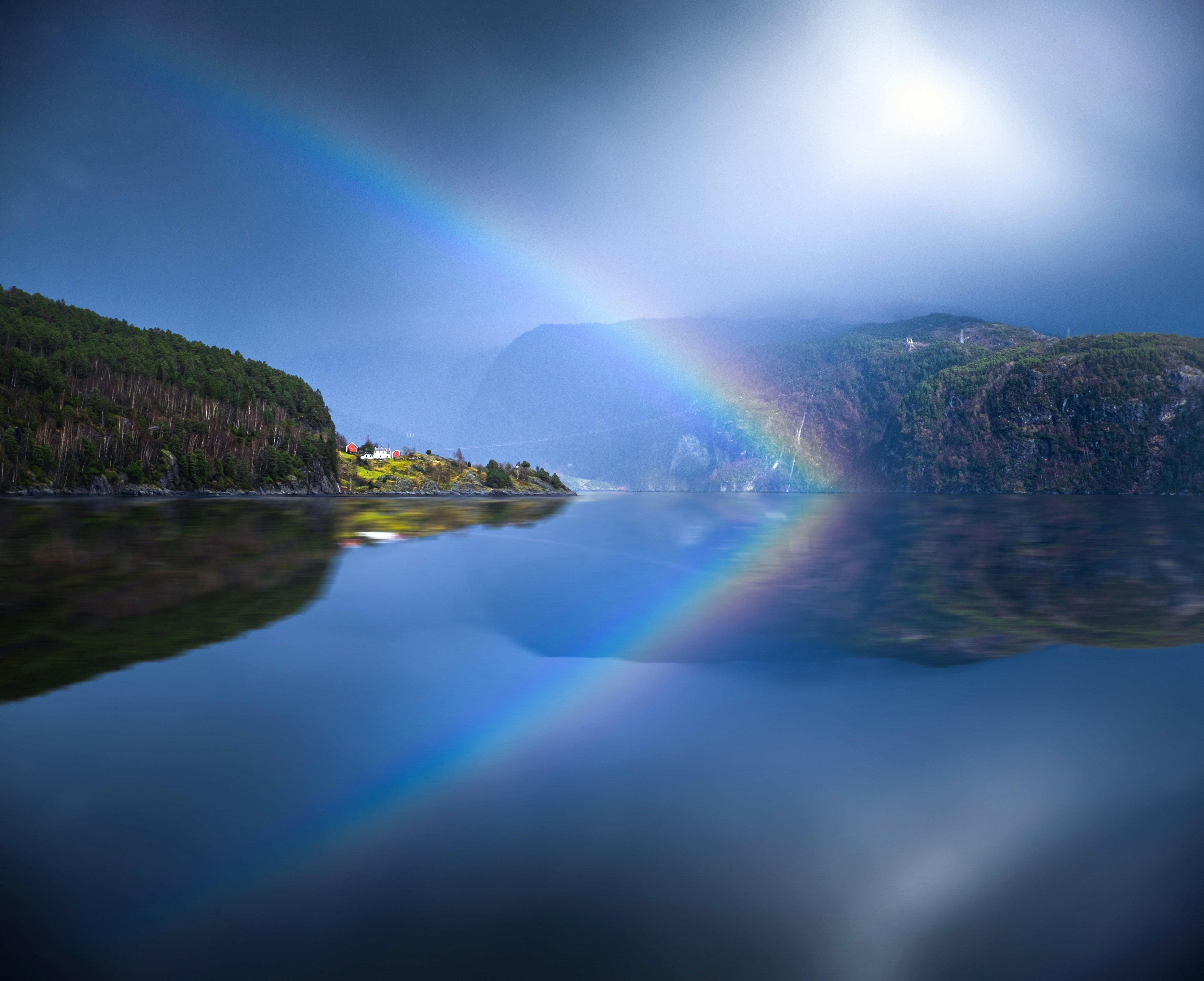 A vibrant rainbow arching over a calm fjord with forested hills and small houses
