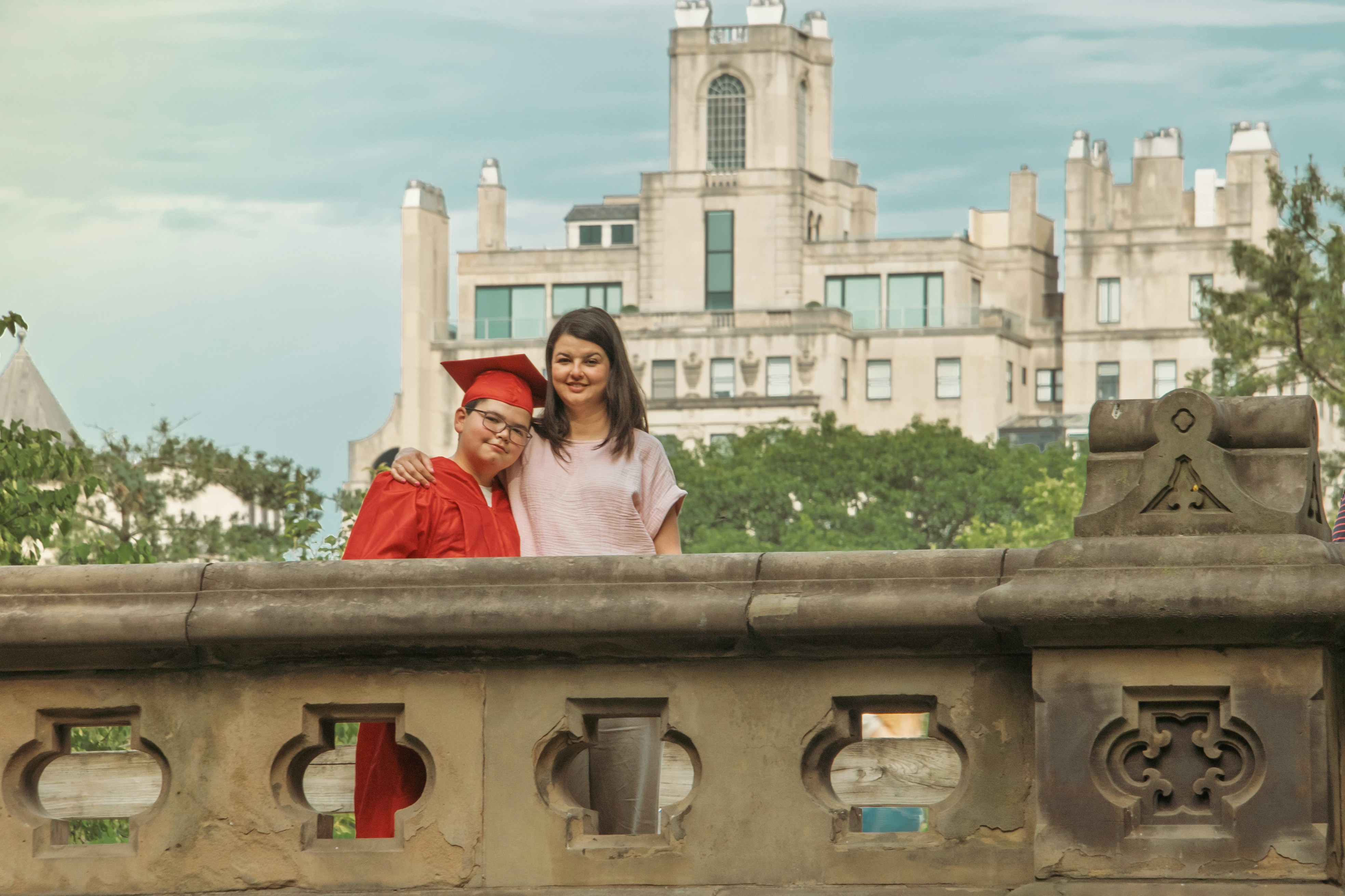 A graduate in a red cap and gown with a woman on a stone bridge