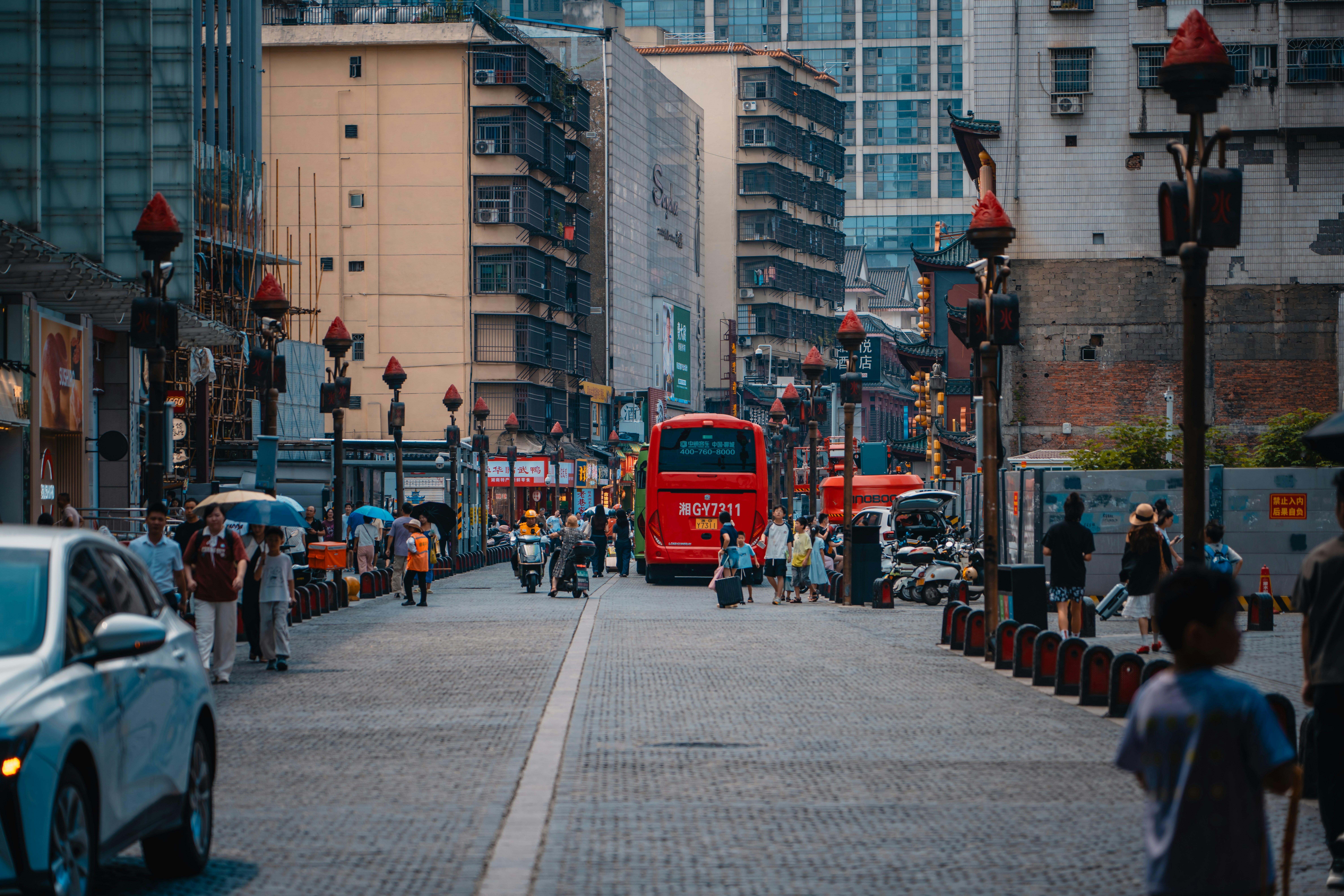 A red bus on a cobblestone street lined with tall buildings and pedestrians