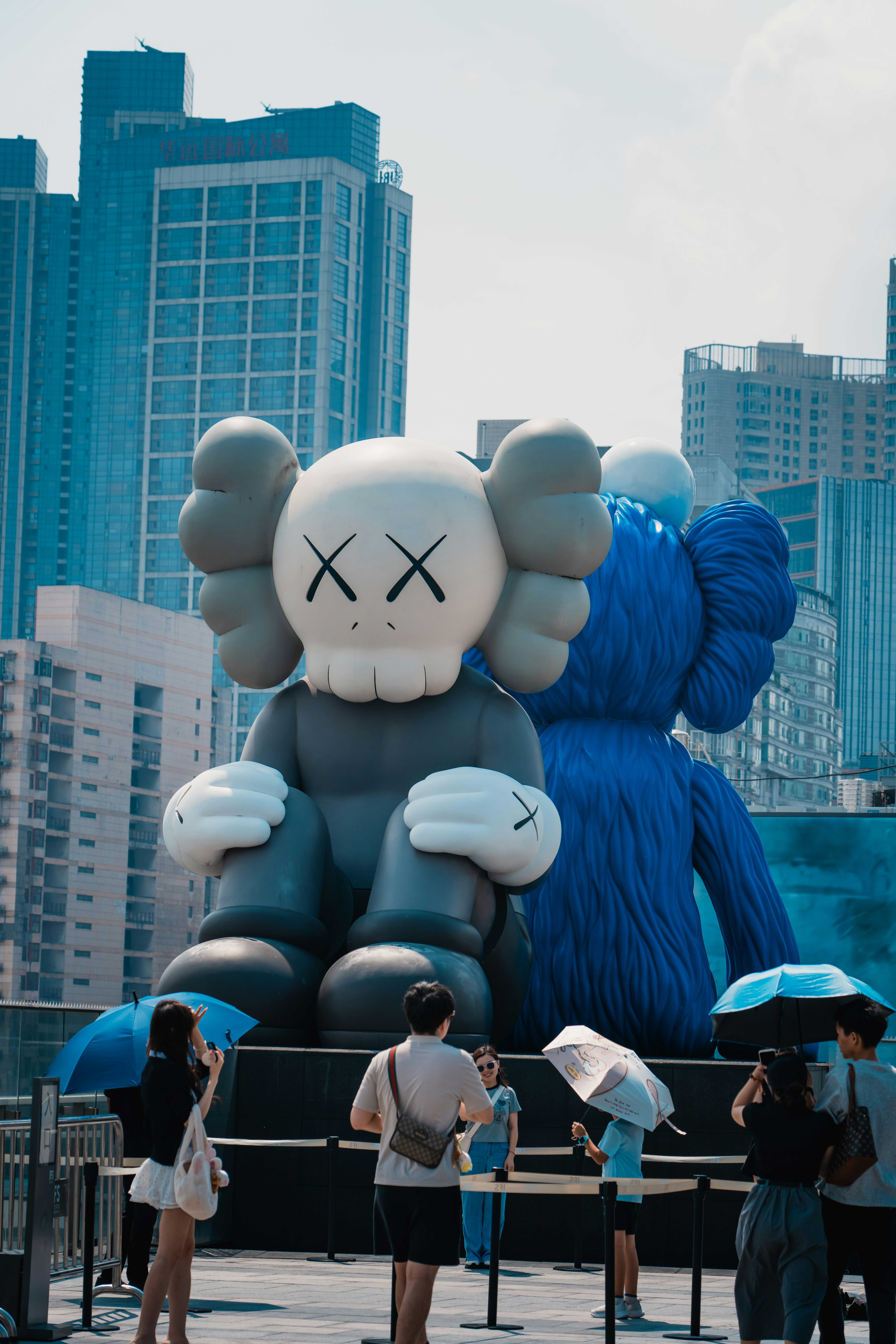 People with umbrellas standing before large grey and blue cartoon character sculptures in a city