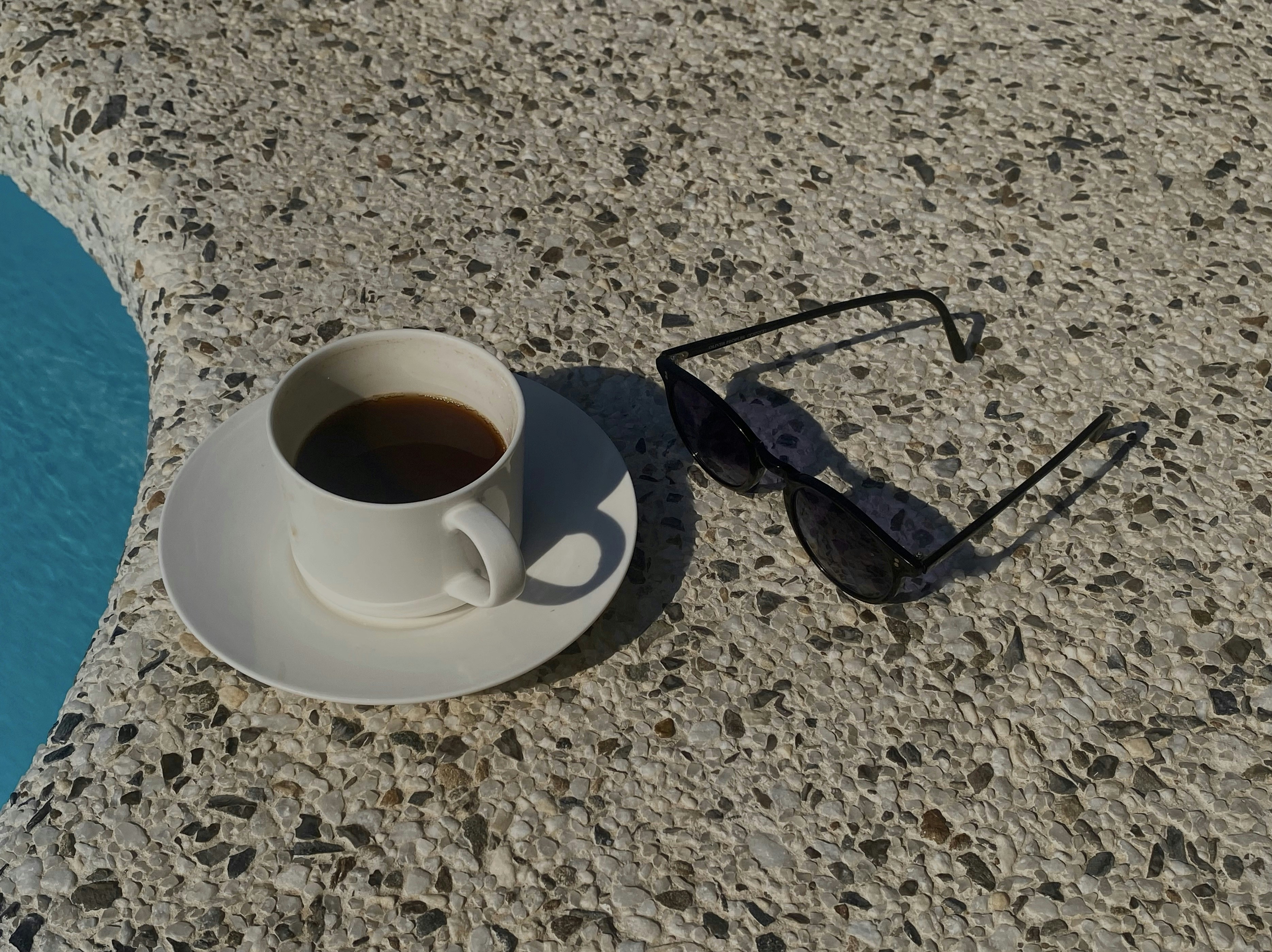 Une tasse de café noir et des lunettes de soleil sur une terrasse de piscine en pierre texturée