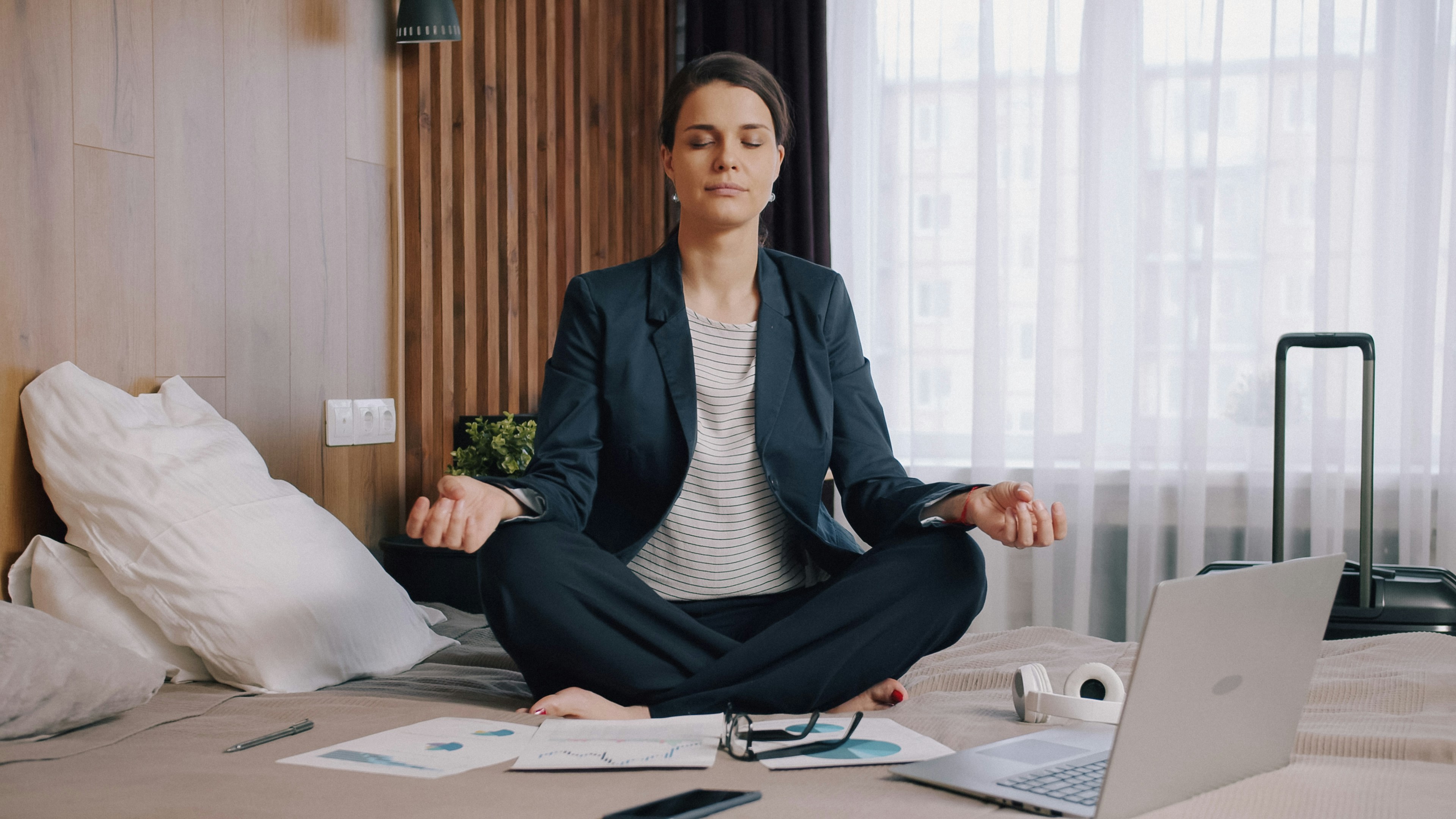 A woman in a business suit meditating on a hotel bed with documents