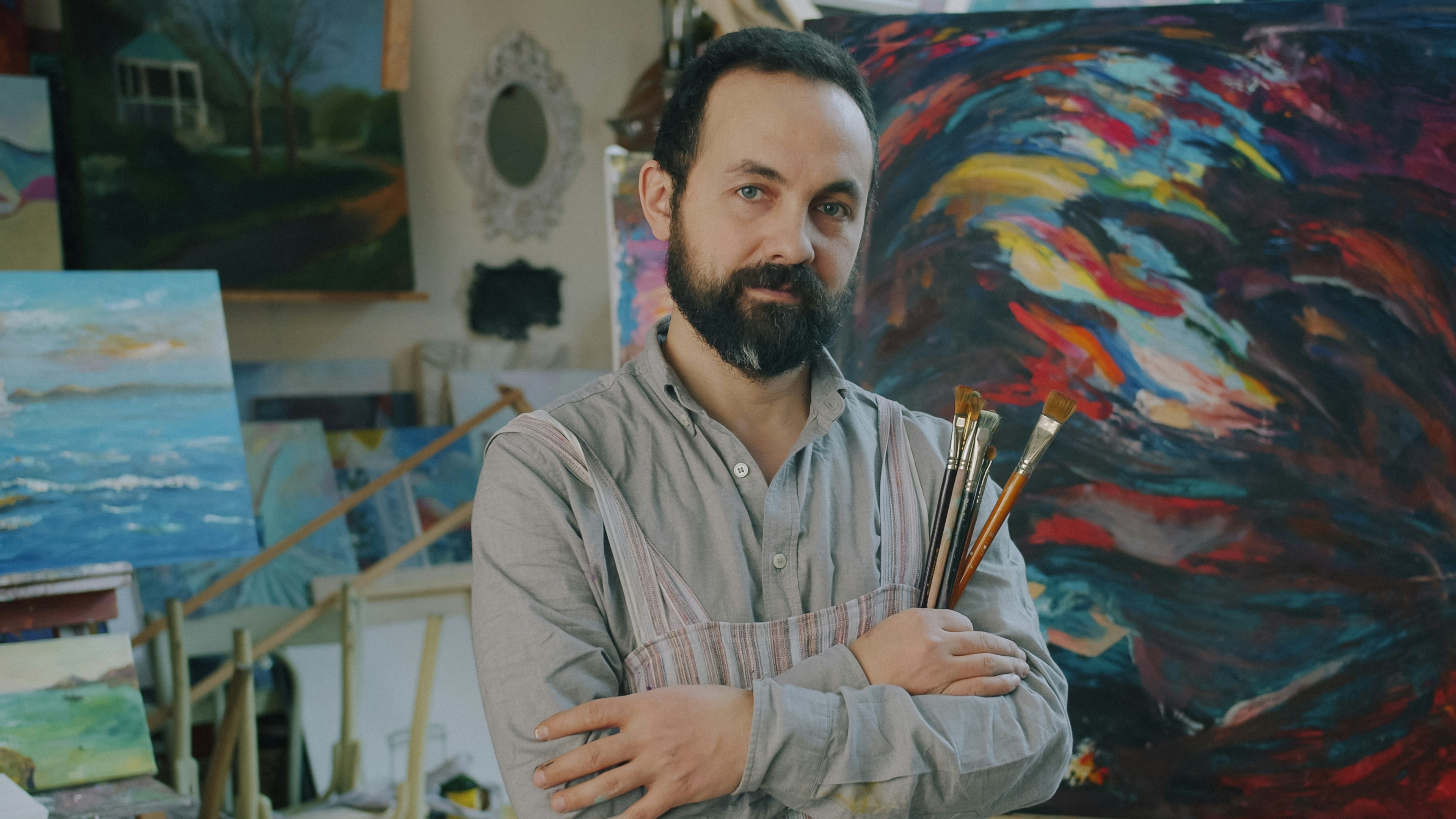 Un hombre con barba sosteniendo pinceles en un estudio de arte con pinturas coloridas