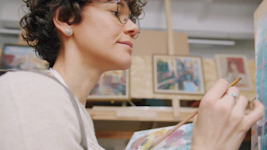A woman with short curly hair and glasses painting on a canvas in a studio