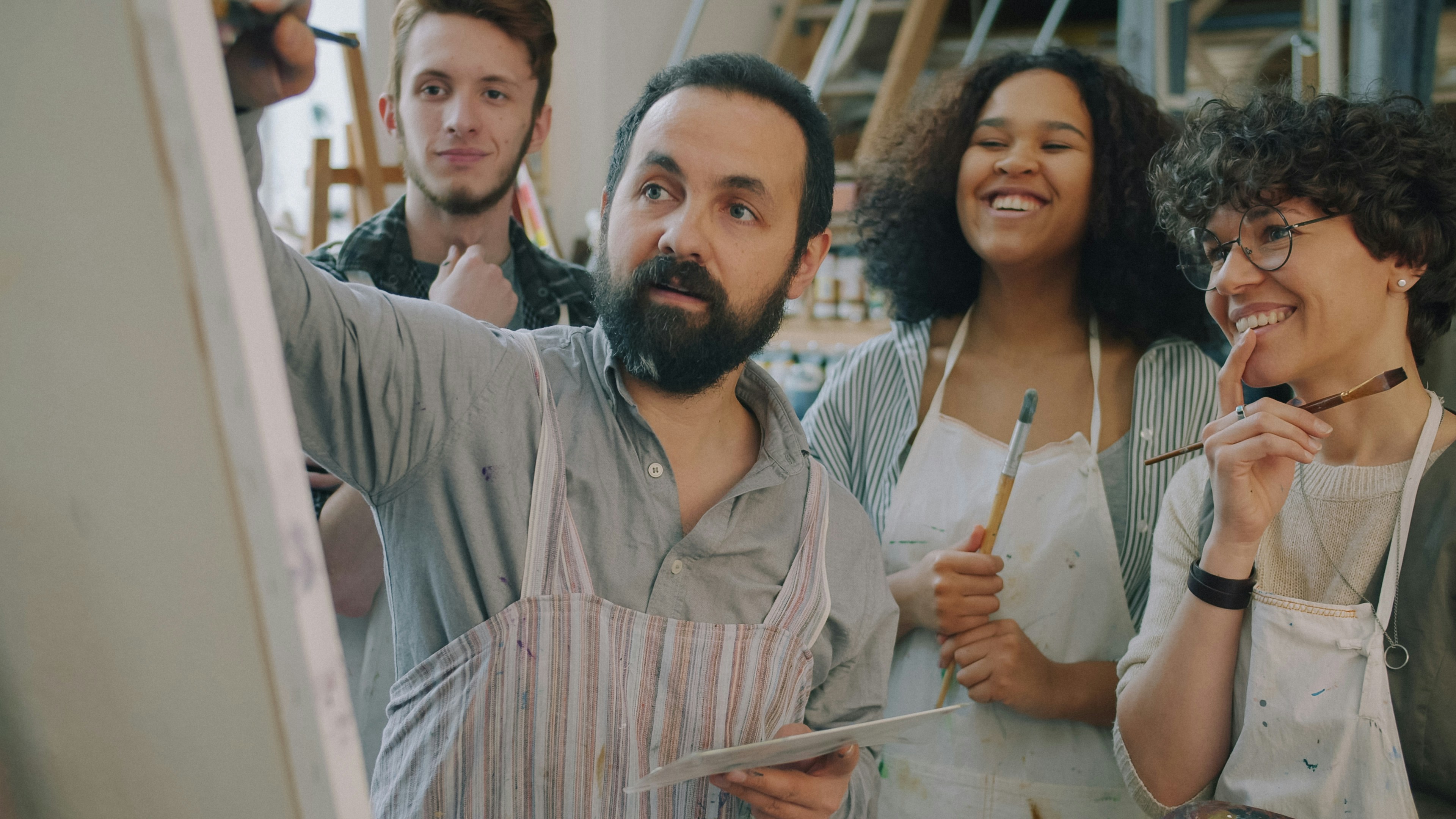 A bearded man painting on a canvas while three students watch in an art studio