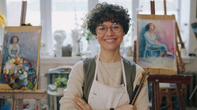 A woman with curly hair and glasses wearing an apron in an art studio