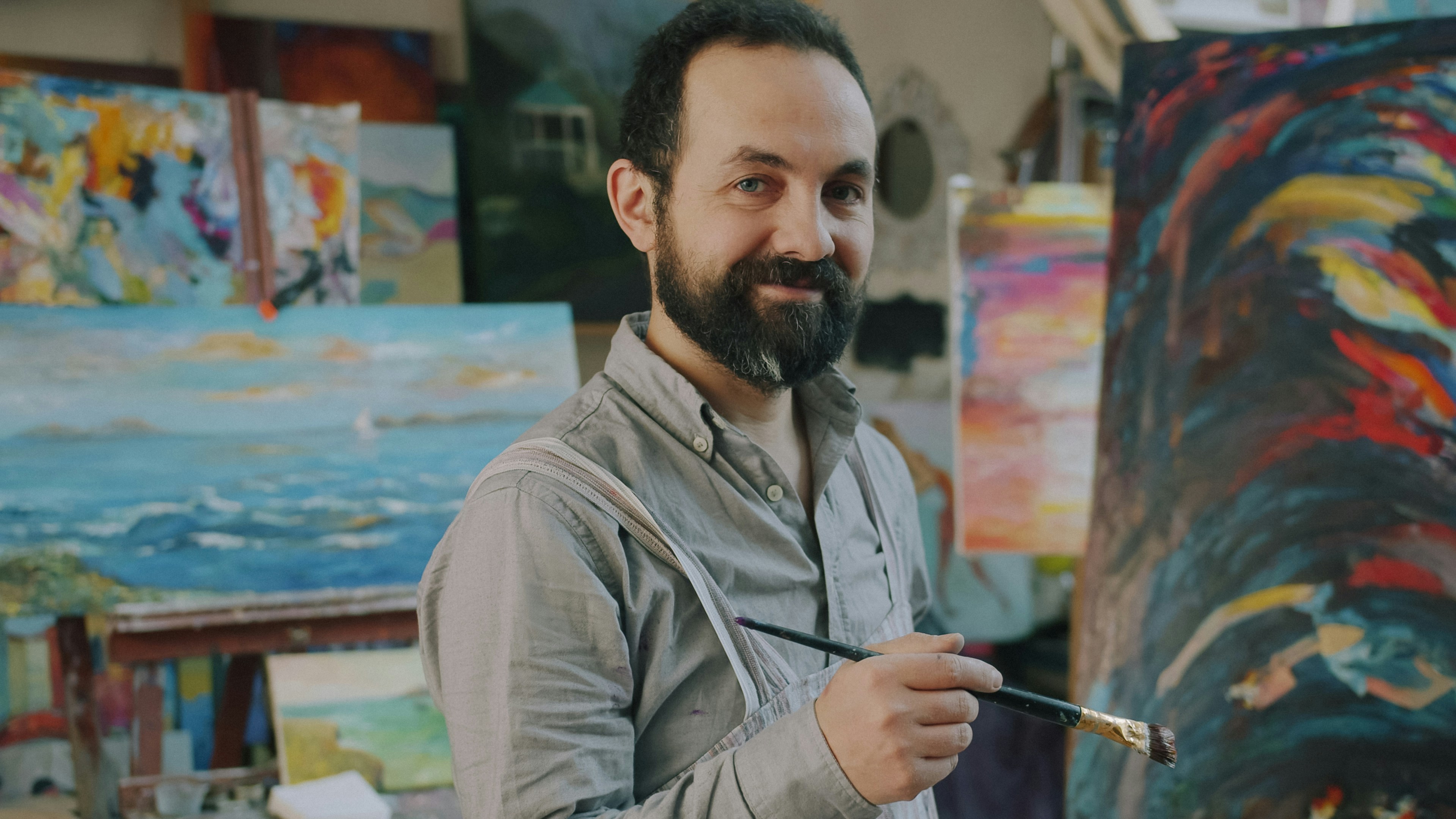 A bearded man holding a paintbrush in a studio filled with colorful paintings