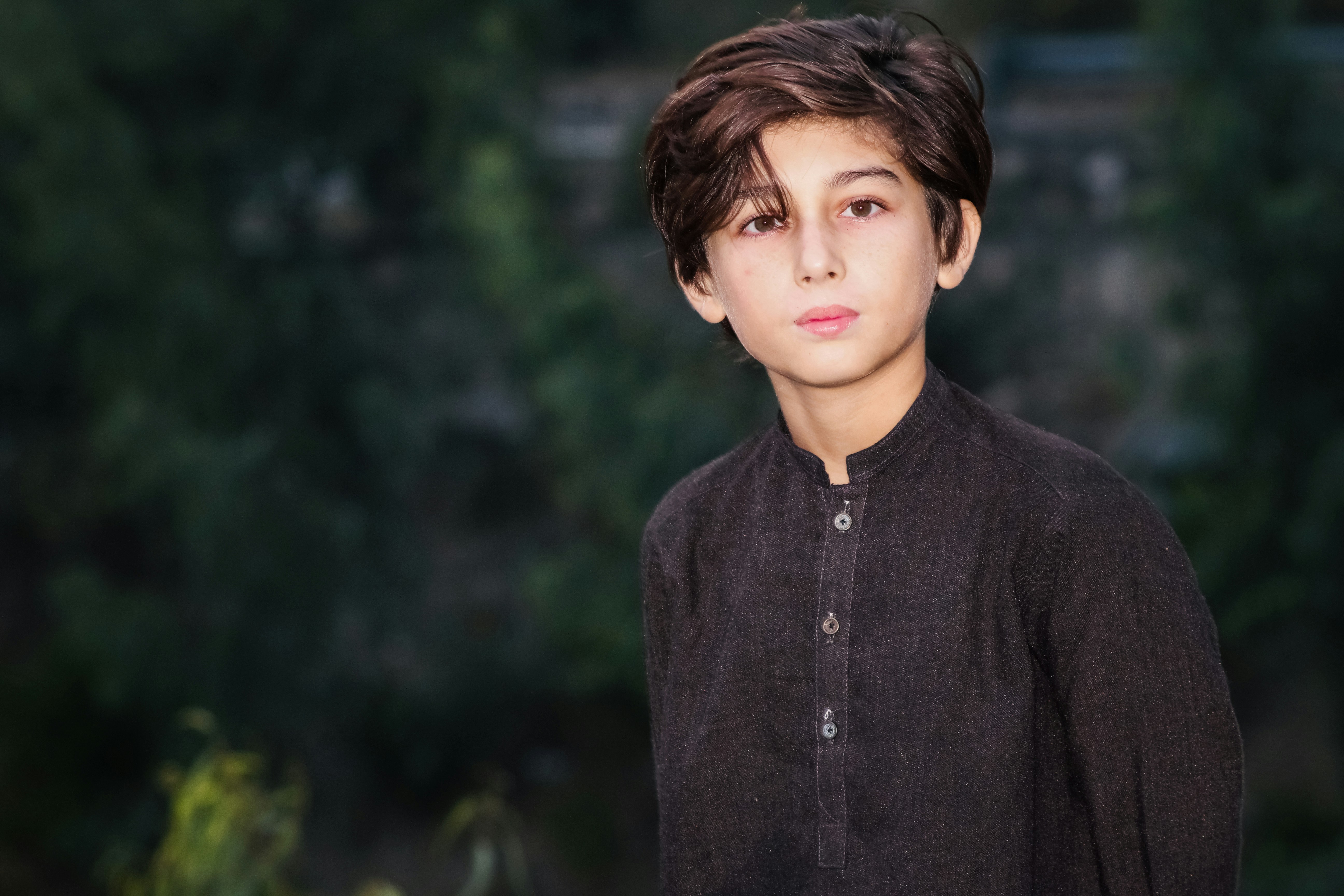 A child with dark hair wearing a dark brown buttoned shirt outdoors