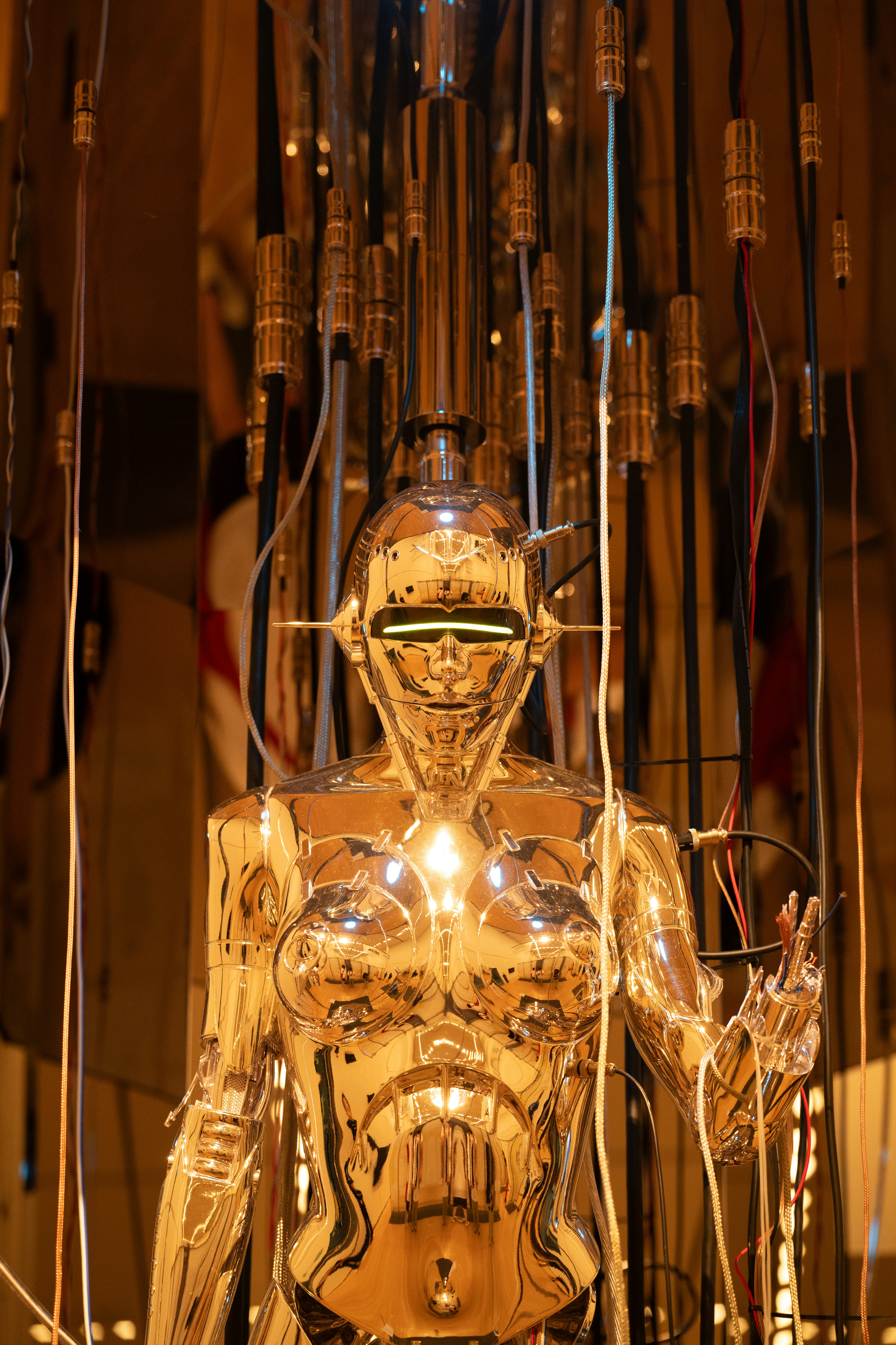 Um robô humanoide dourado brilhante com olhos brilhantes cercado por cabos elétricos pendurados