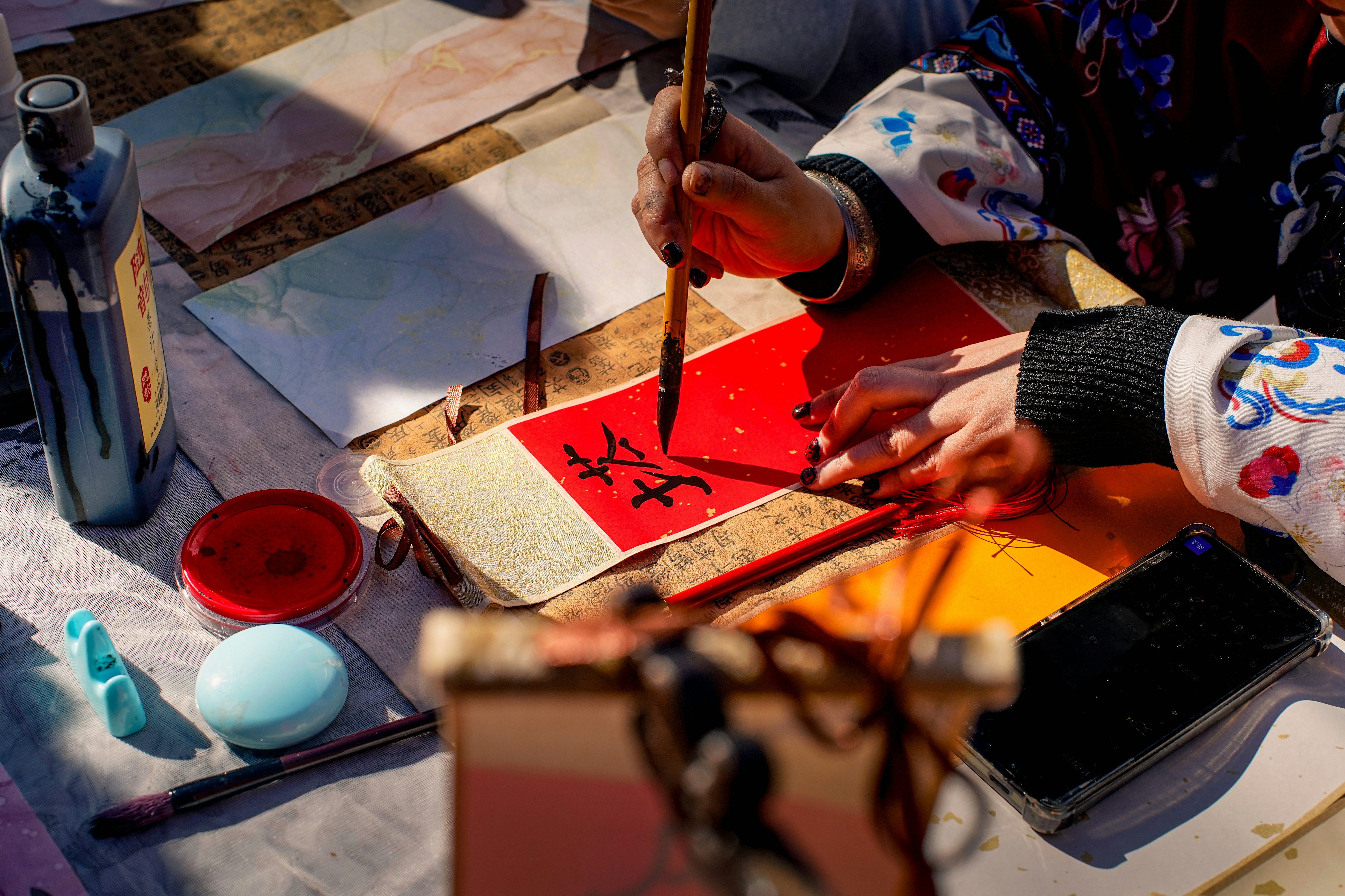 A person writing Chinese calligraphy with a brush on red paper at a table