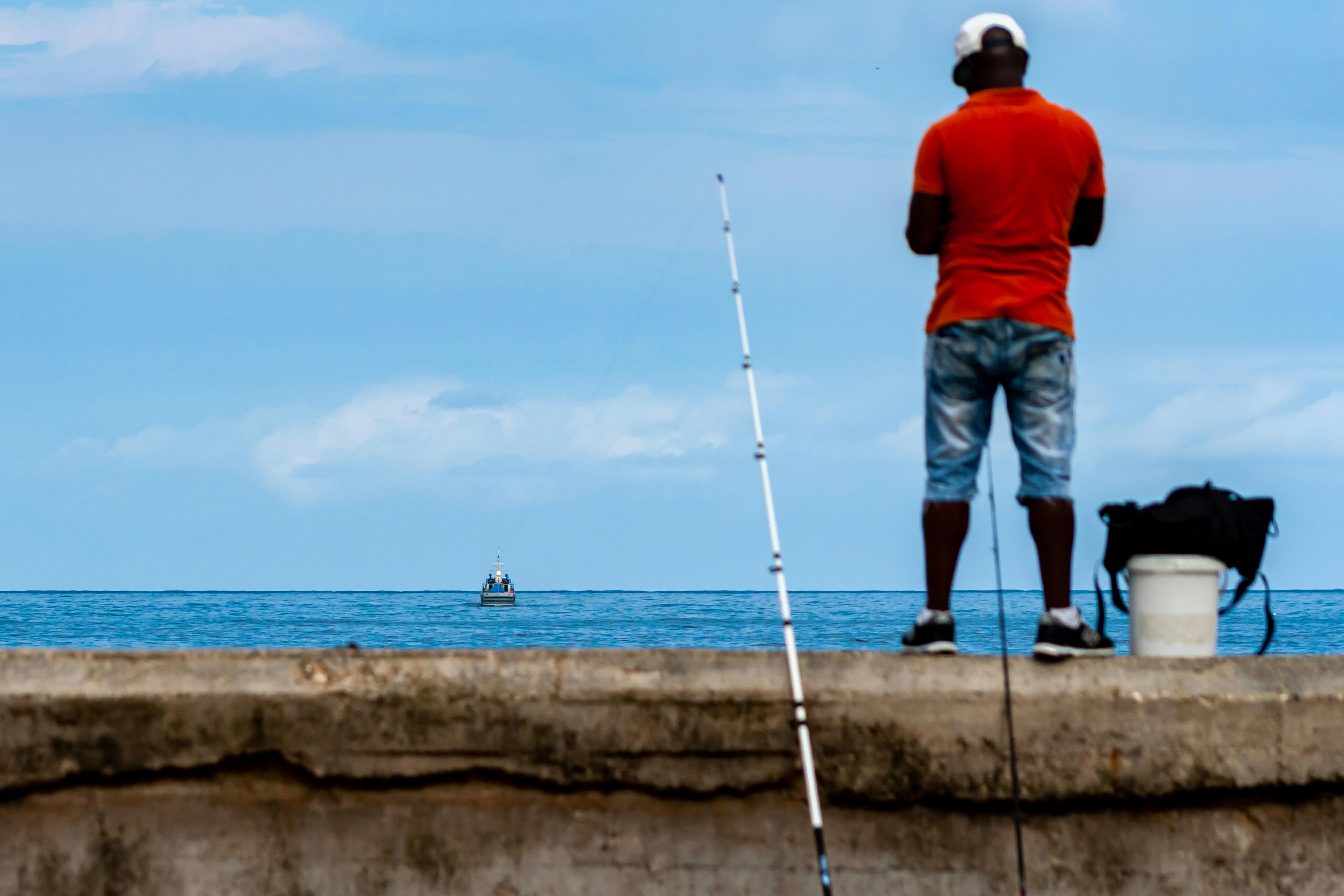 A person in an orange shirt fishing from a concrete pier toward a boat