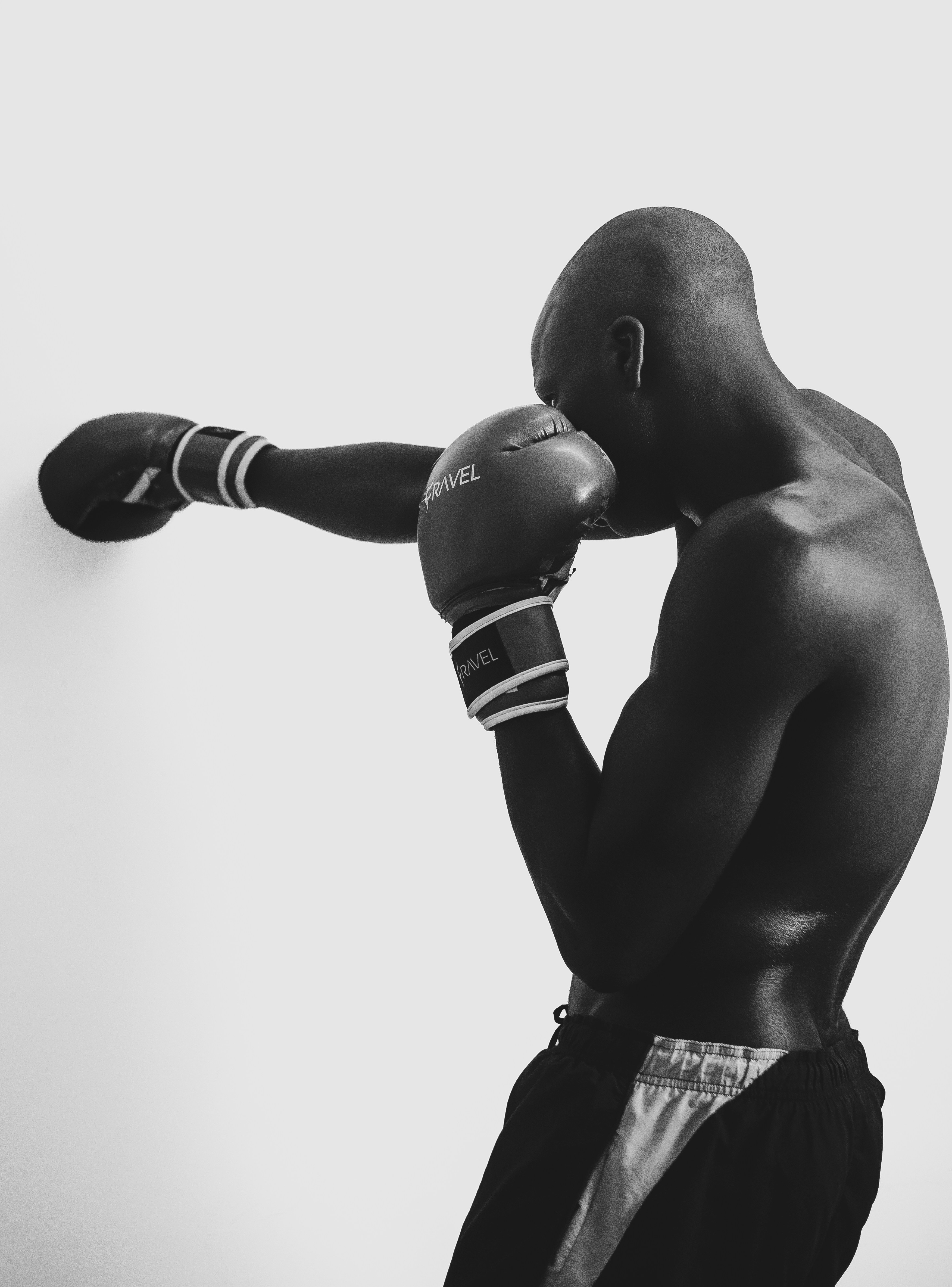A shirtless man wearing boxing gloves punching against a plain white wall