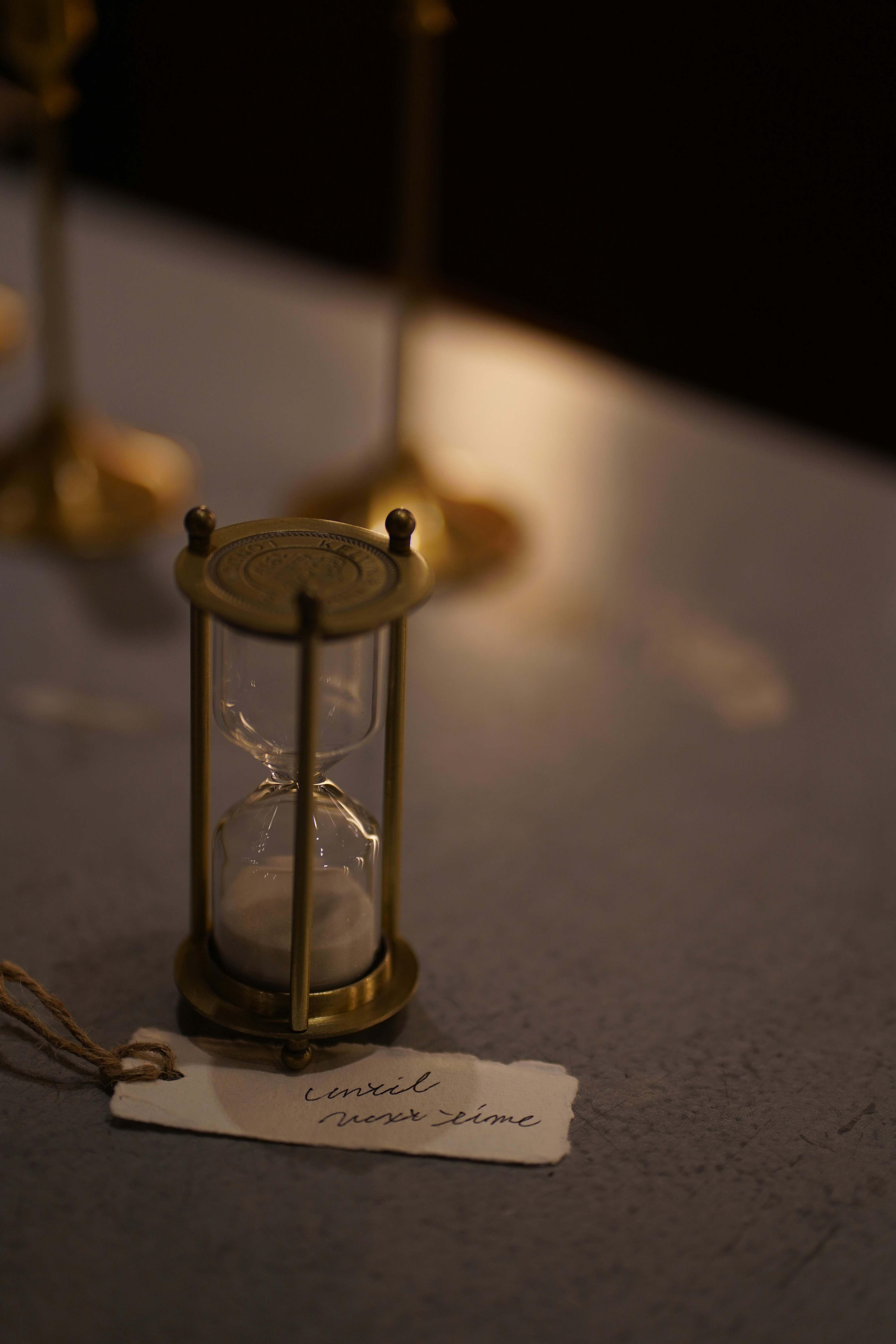 A brass hourglass with white sand beside a small paper tag on a table